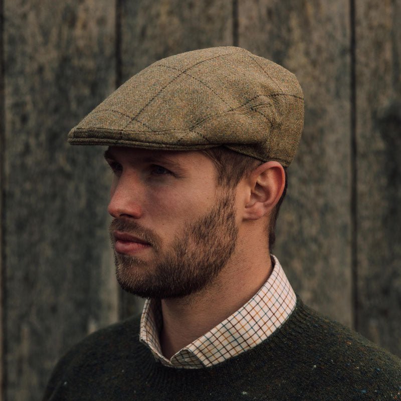 A bearded man wears the Auld Stock Conon Cap by Campbell's of Beauly, paired with a checked button-up shirt and dark sweater, as he stands before a weathered wooden wall, gazing to the left.