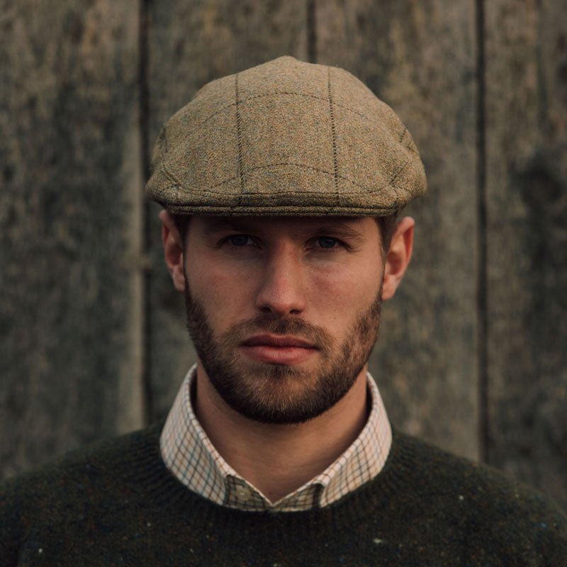 A man with a short beard and serious expression wears the Campbell's of Beauly Auld Stock Conon Cap, a plaid shirt, and a dark sweater, standing before a weathered wooden background.