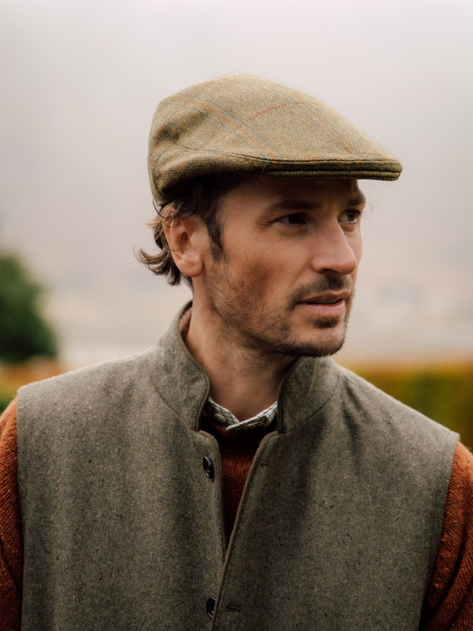 A man wears the Auld Stock Conon Cap by Campbell's of Beauly, paired with a grey vest, rust sweater, and collared shirt. He stands outdoors against a softly blurred natural background, gazing to the side neutrally.