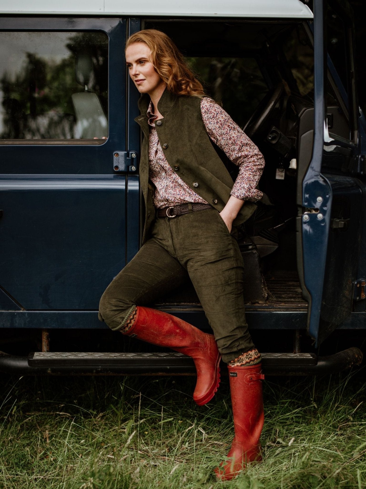 A person with long red hair in a floral shirt, green vest, and red rubber boots stands on grass by an open blue SUV door, wearing Campbell's of Beauly Corduroy Plus Twos trousers.
