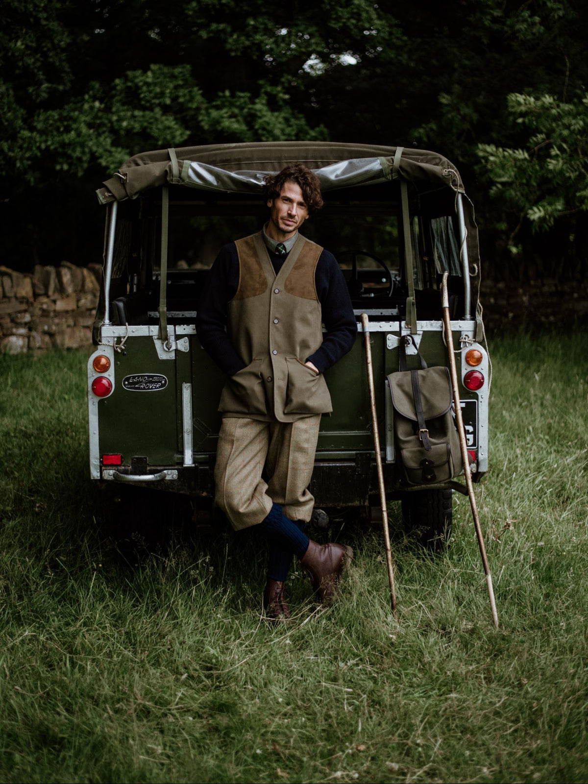 A man in country attire, wearing a Campbell's of Beauly Cotton Shooting Waistcoat, leans against a vintage green Land Rover on grass, with walking sticks and a satchel—ideal for countryside pursuits amid trees and a stone wall.