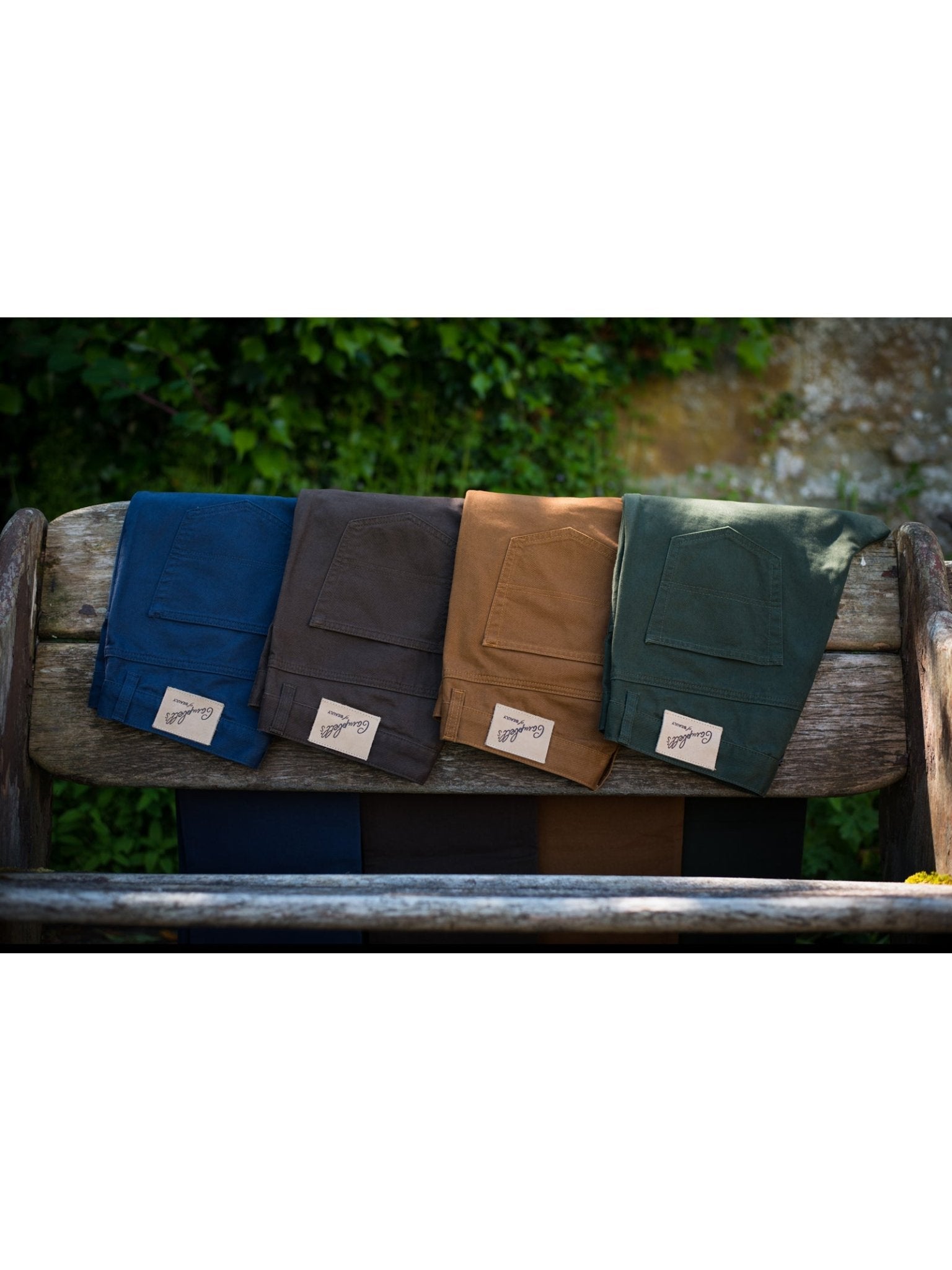 Four pairs of Campbell's of Beauly Colour Cotton Jeans in navy, dark brown, tan, and dark green—each with a classic five-pocket design—are neatly arranged on a rustic wooden bench outdoors against green foliage.