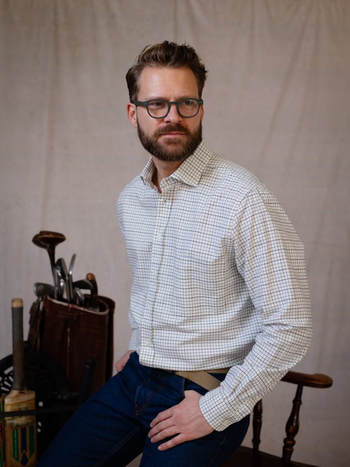 A man with glasses and a beard, wearing the Campbell’s of Beauly Windowpane Shirt in brushed cotton and jeans, sits on a wooden chair. Vintage golf clubs in a leather bag are positioned behind him, set against a plain light-colored backdrop.