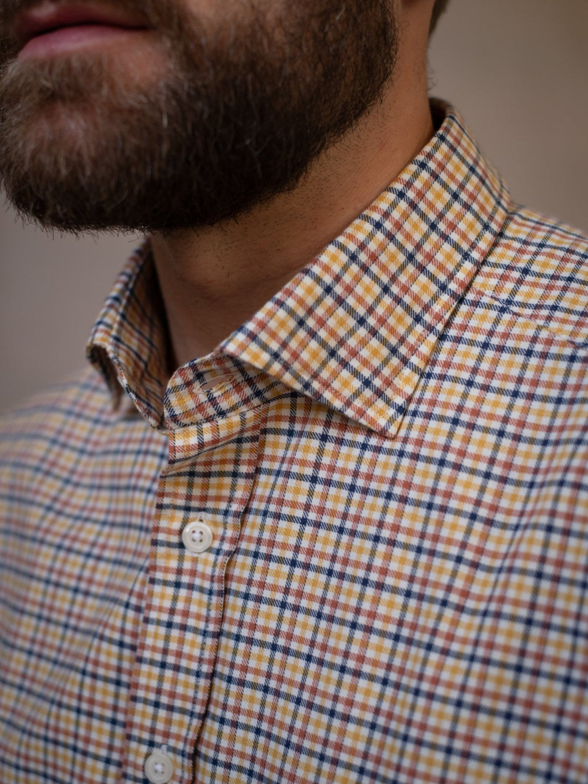 A close-up of a man’s lower face and neck, featuring a brown beard and the buttoned-up Windowpane Shirt by Campbell’s of Beauly, distinguished by its yellow, brown, and blue plaid pattern in soft brushed cotton.