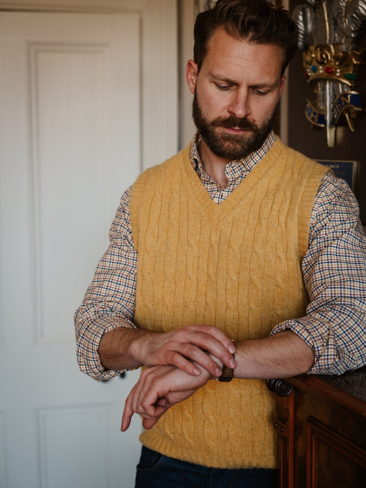 A man with a beard, dressed in a yellow sweater vest over a Campbell’s of Beauly Windowpane Shirt, stands indoors and gazes thoughtfully down at his wristwatch.