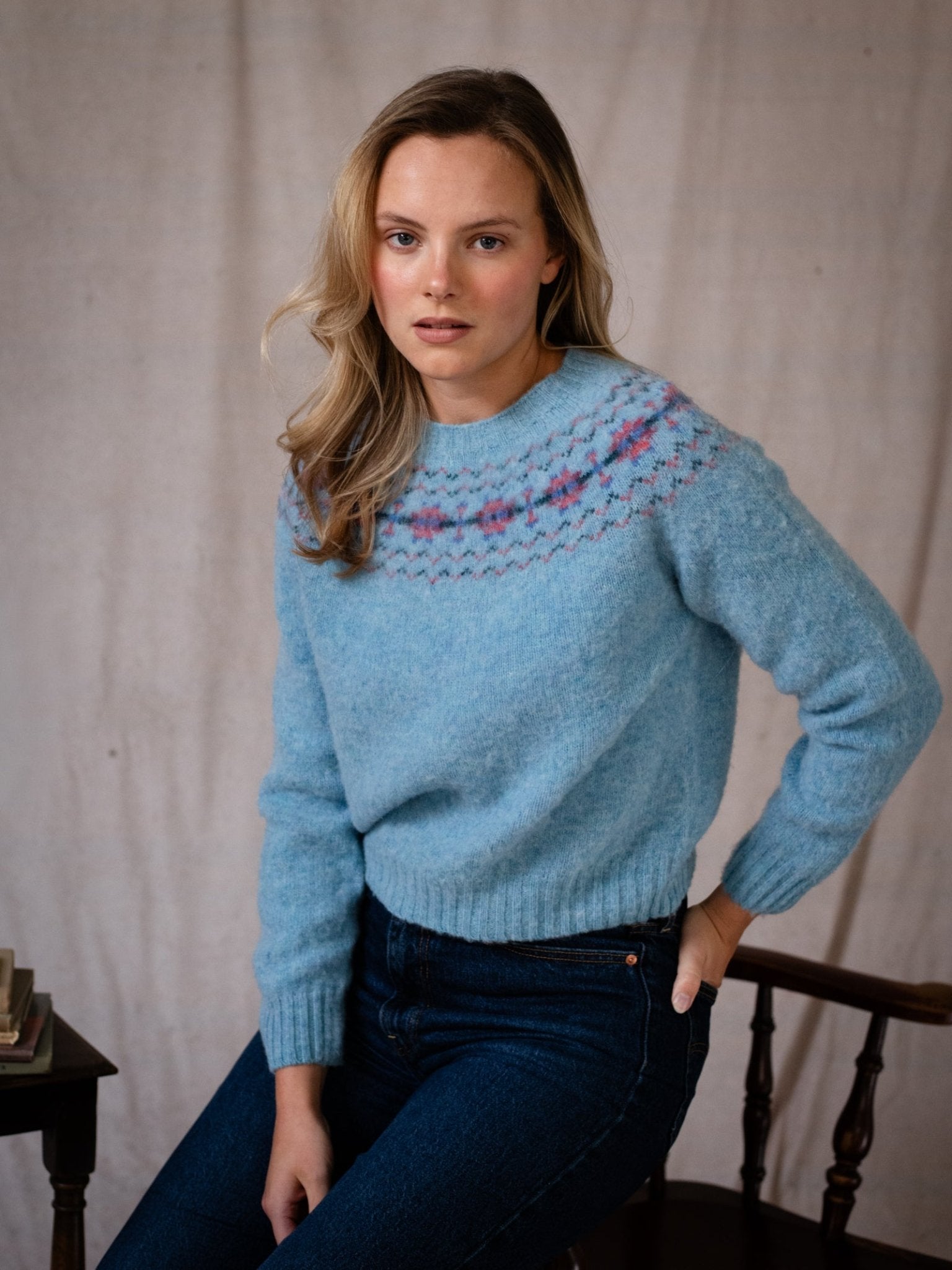 A woman with long blonde hair sits on a wooden chair, wearing the Campbell's of Beauly Shaggy Cropped Fairisle Crew in Dewdrop and dark blue jeans, looking at the camera with a neutral expression.