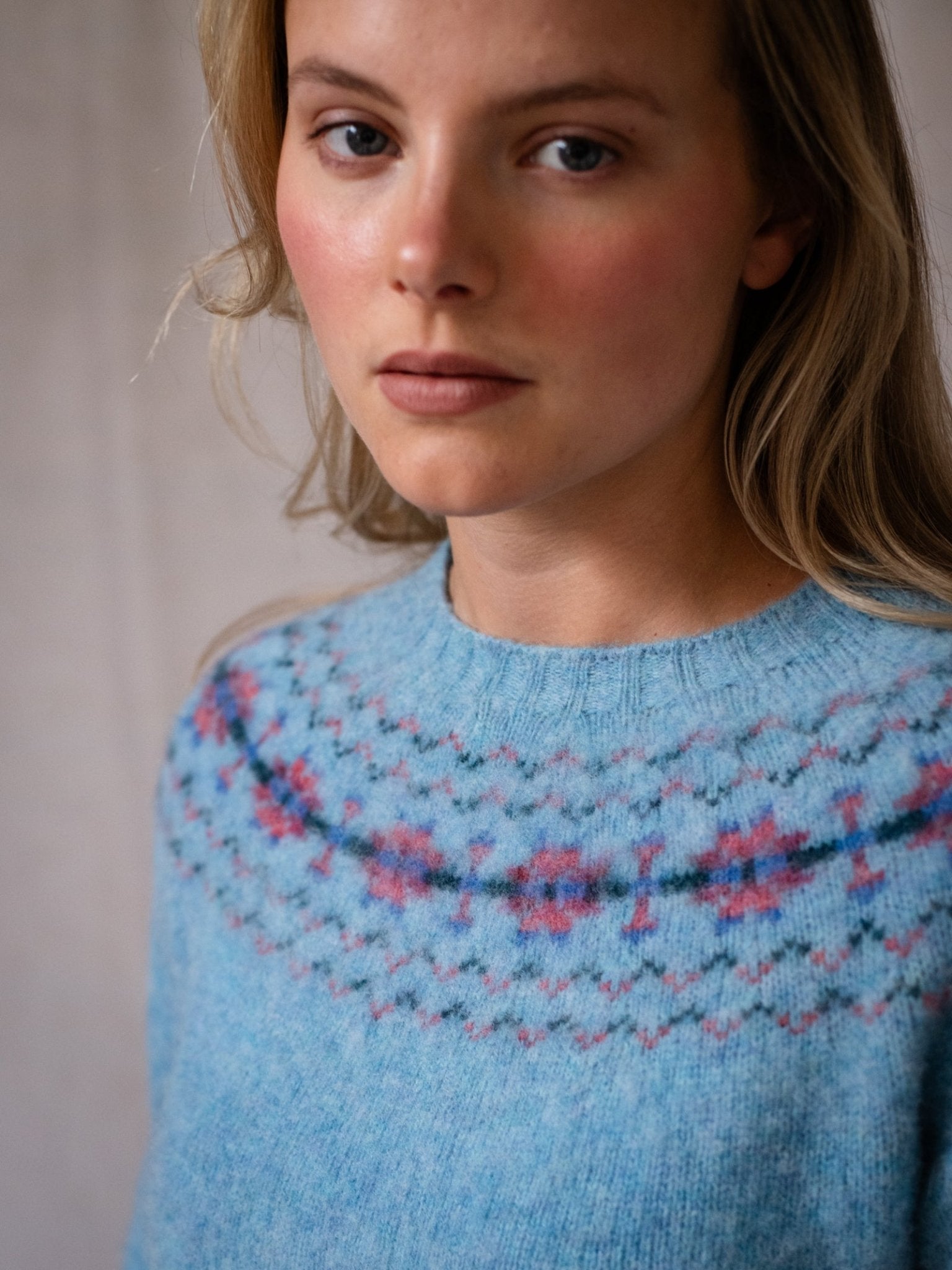 A person with light skin and long blonde hair wears the Campbell's of Beauly Shaggy Cropped Fairisle Crew, a light blue Pure New Wool sweater with a pink and blue patterned yoke. The background is neutral and out of focus.