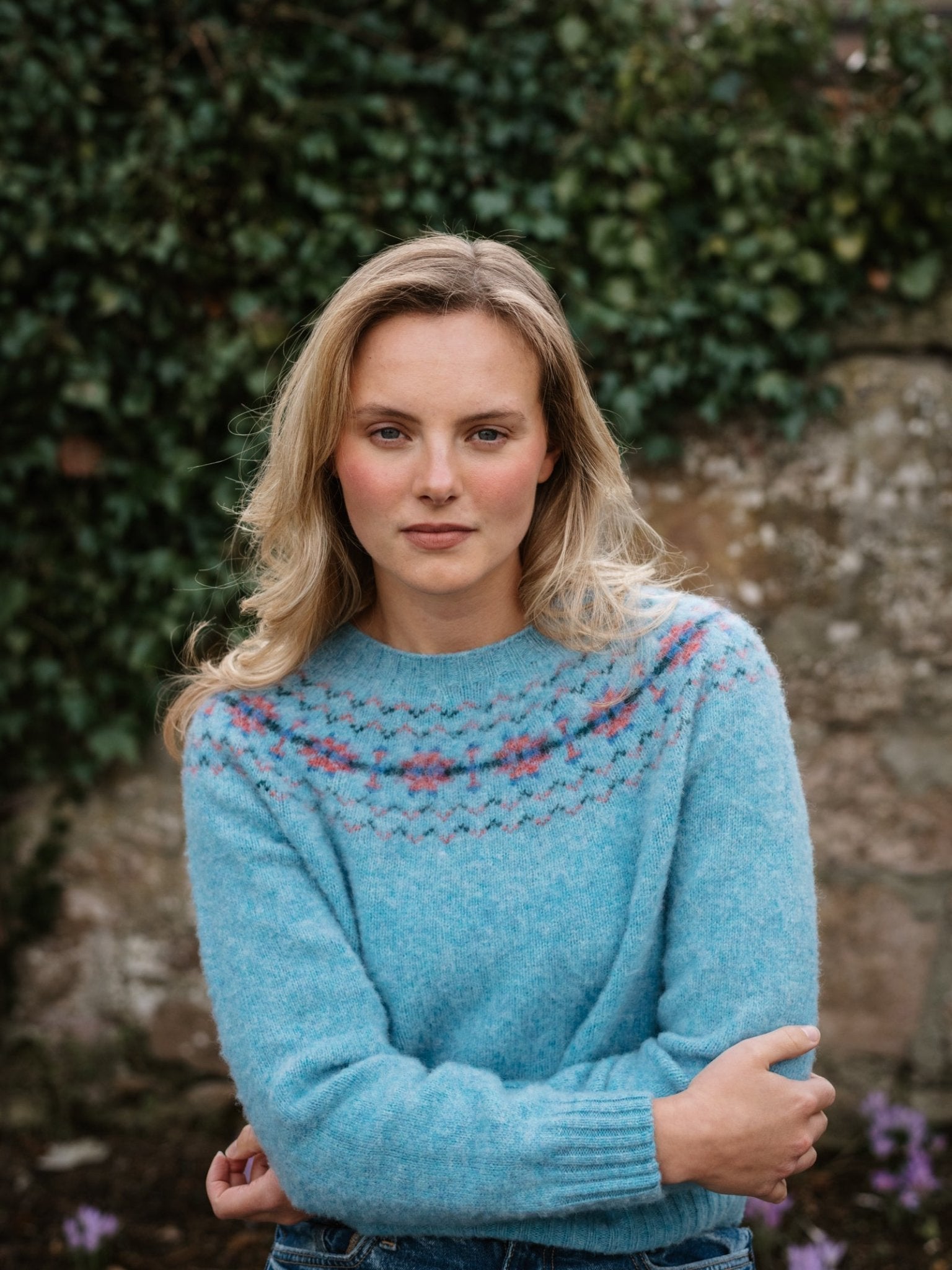A woman with blonde hair wears the Shaggy Cropped Fairisle Crew by Campbell's of Beauly, standing outdoors in front of a green ivy-covered stone wall and looking calmly at the camera.
