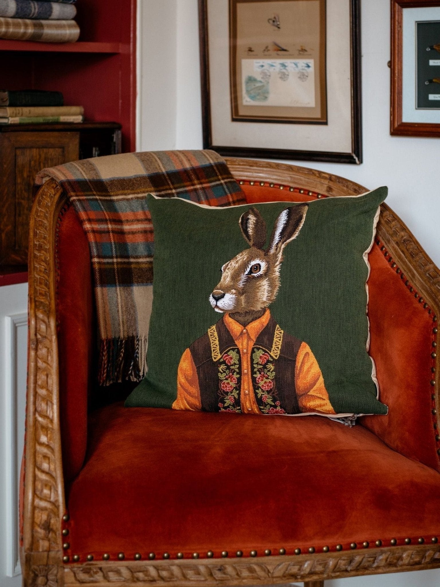 A cozy room with framed art on the wall features a red velvet armchair adorned with a plaid blanket and the Campbell’s of Beauly Alpine Hare Cushion Cover, which showcases an alpine hare dressed in embroidered clothing.