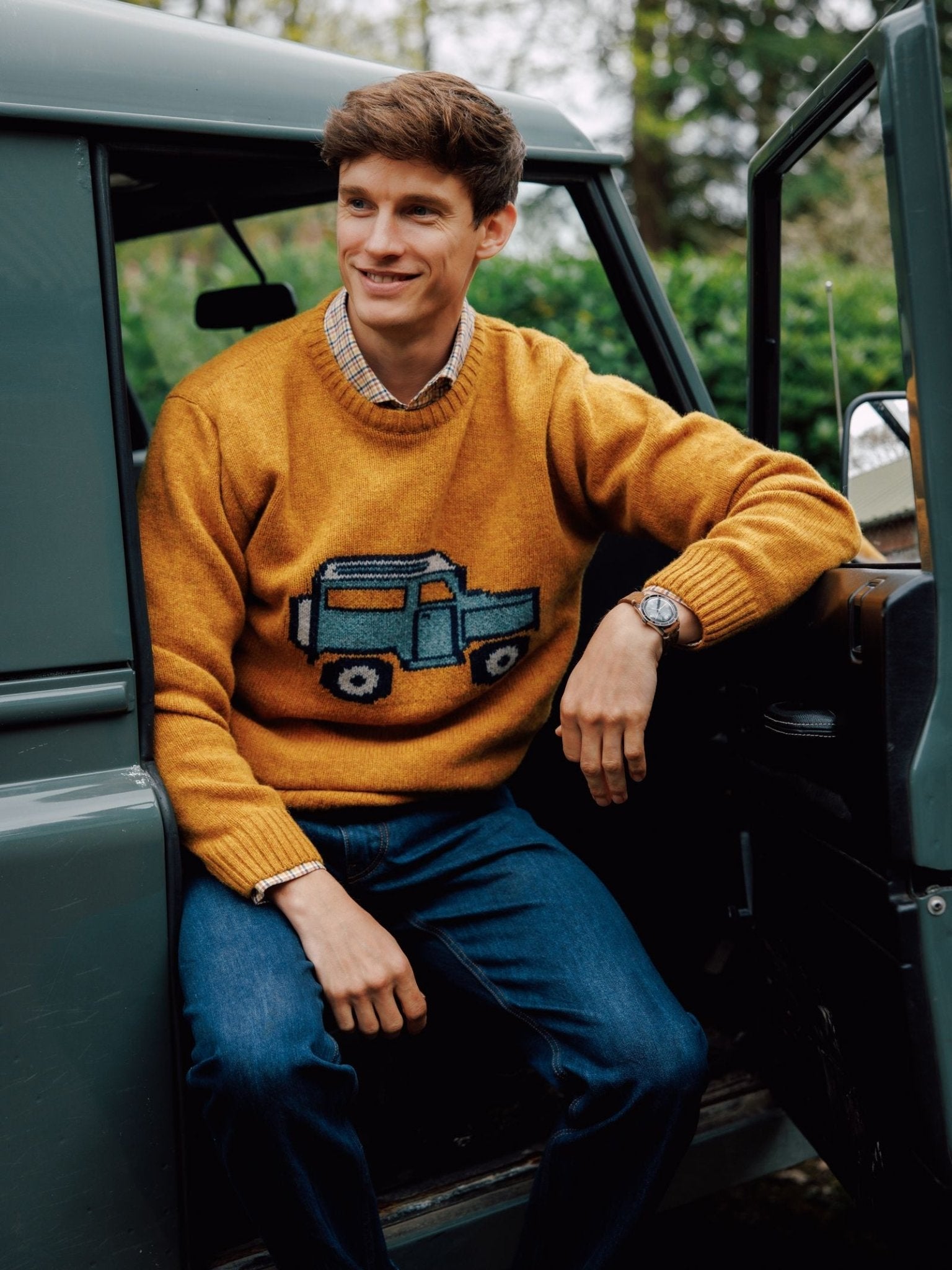 A young man with short brown hair, wearing the Campbell's of Beauly Unisex Land Rover Defender Jumper, blue jeans, and a watch, smiles while sitting in the open door of a green vehicle with trees in the background.