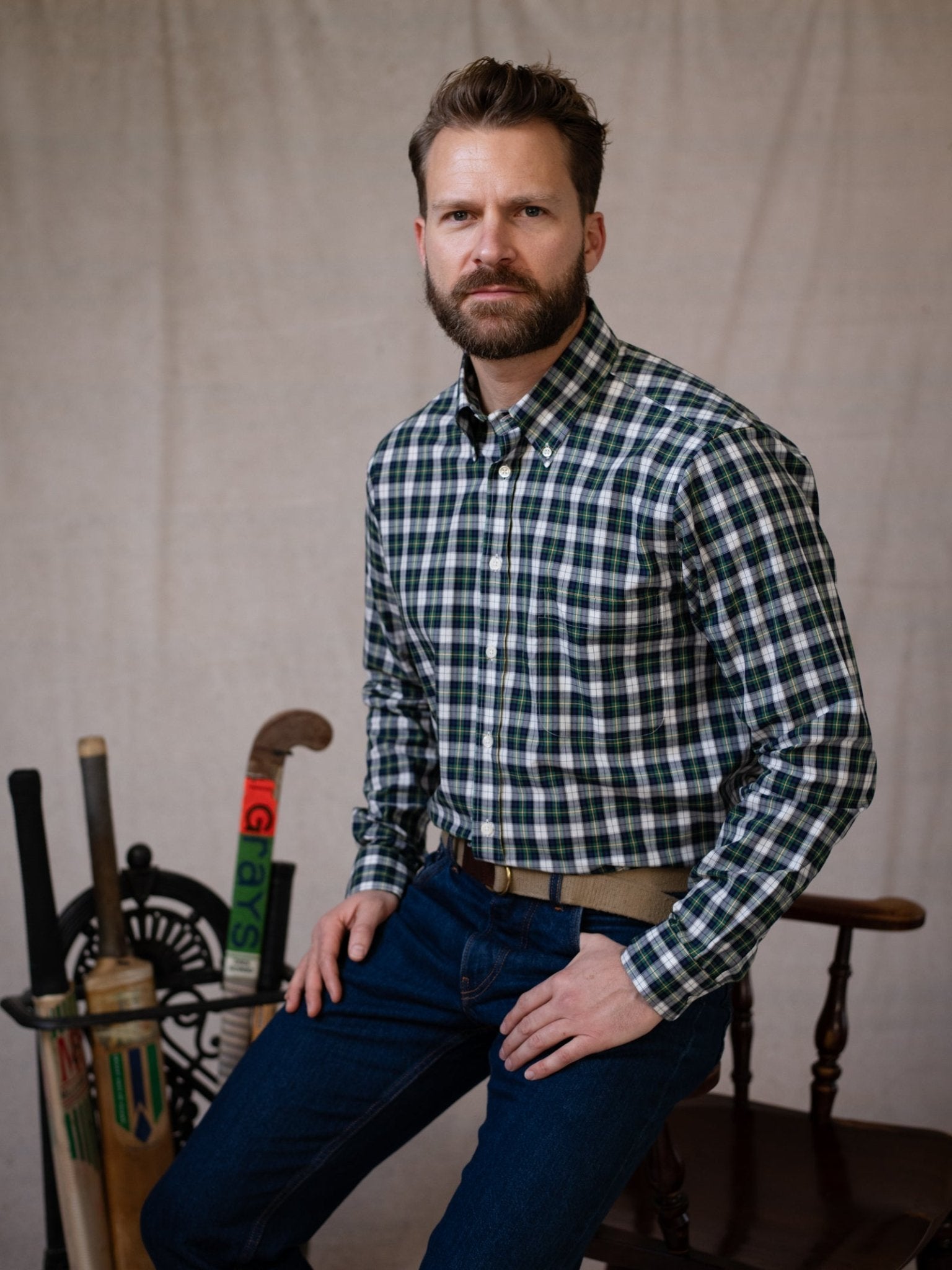 A bearded man wearing Campbell's of Beauly Cotton Tartan Button Down Shirt in green and white sits on a wooden chair, with cricket bats, a field hockey stick, and a wheel behind him against a neutral backdrop.