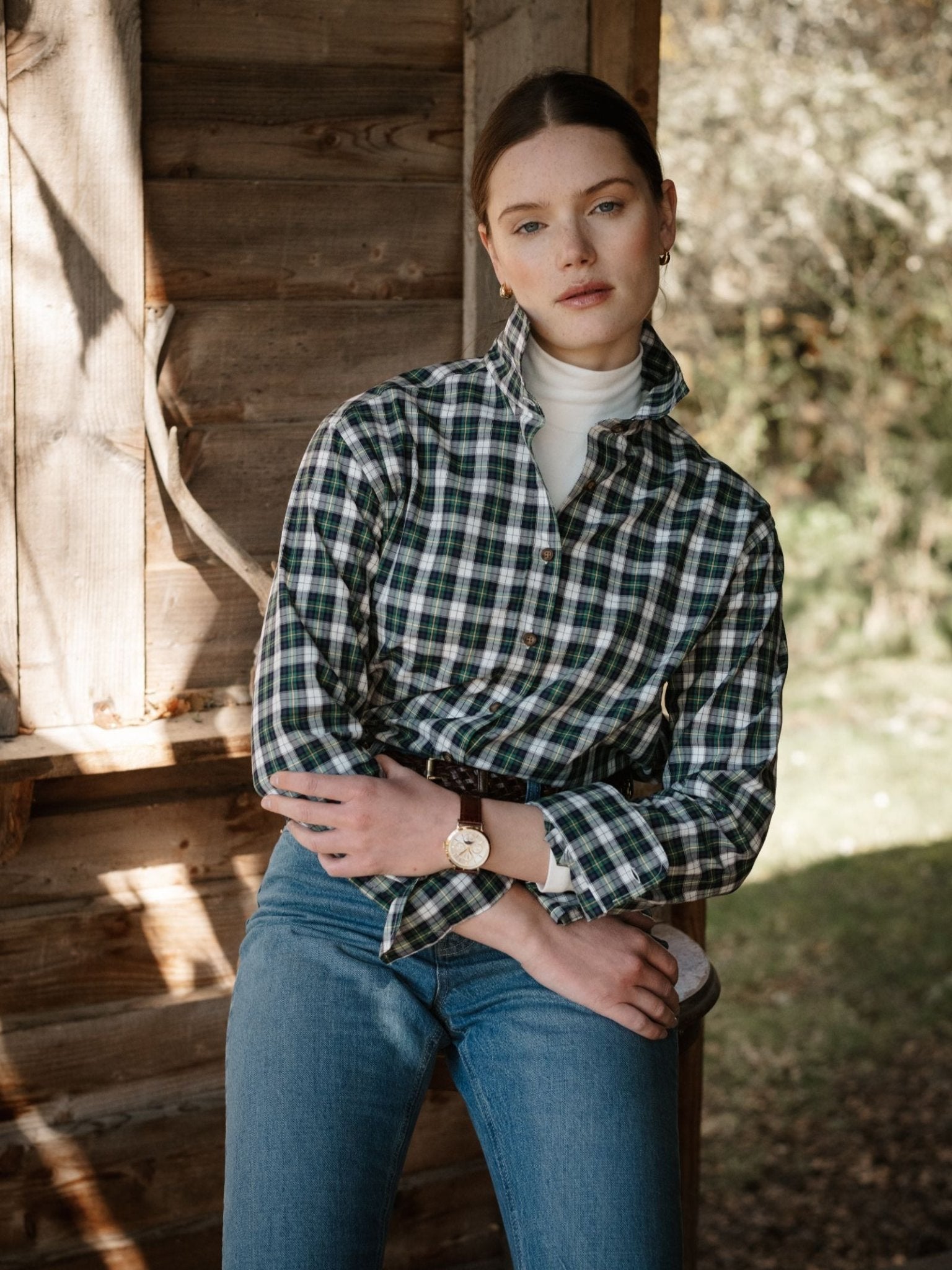 A woman with light skin and brown hair sits casually on a wooden porch, wearing a Campbell's of Beauly Cotton Tartan Shirt over a white turtleneck and blue jeans, surrounded by trees and sunlight in the background.