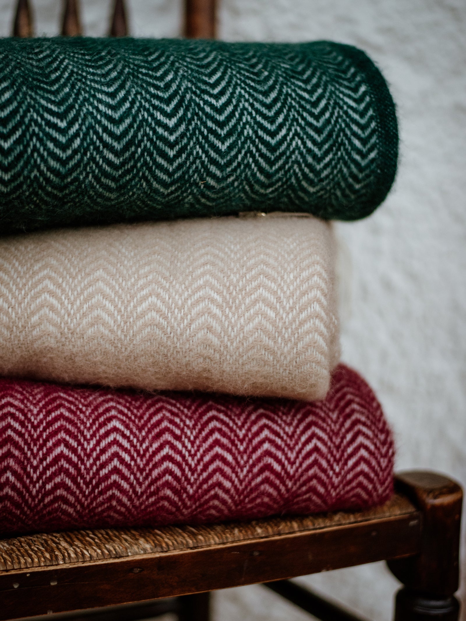 Three folded Campbells of Beauly Dune Throws are stacked on a wooden chair: dark green, beige, and burgundy. Their zigzag design adds a cozy touch to any room against a textured white wall.