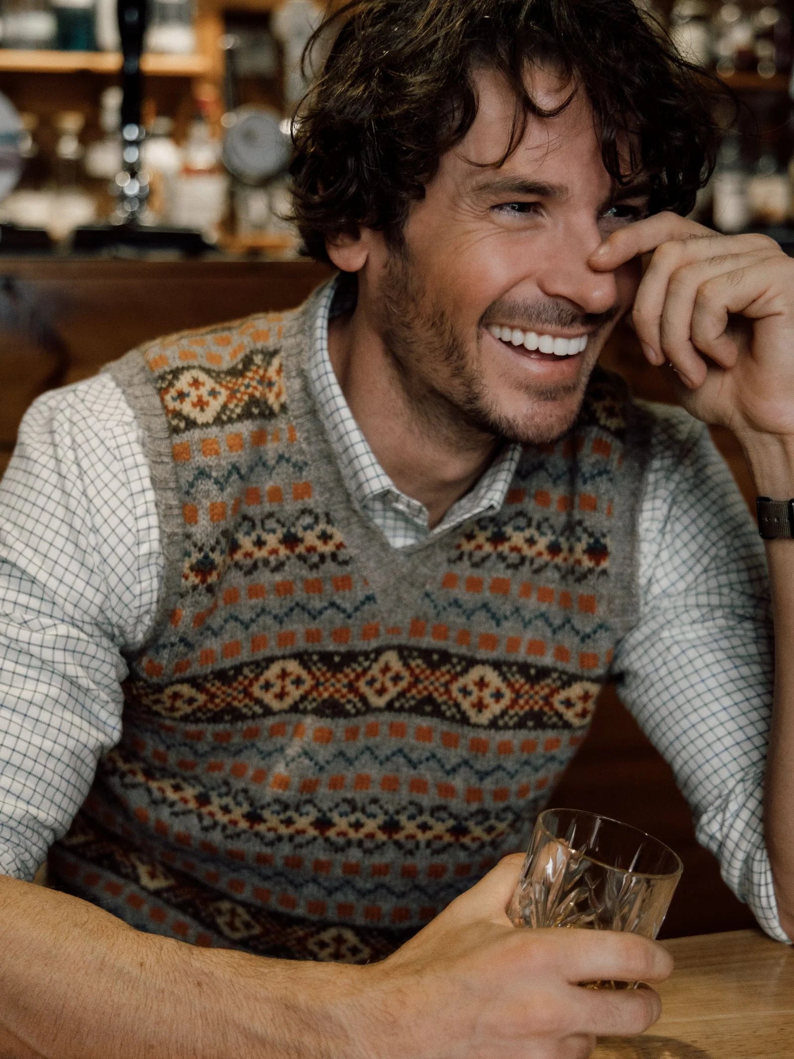 A smiling man with wavy hair sits at a table, wearing the Campbell’s of Beauly Fairisle Slipover—crafted from soft Shetland wool—over a checkered shirt. He holds a glass in one hand and rests his other hand near his face, laughing.