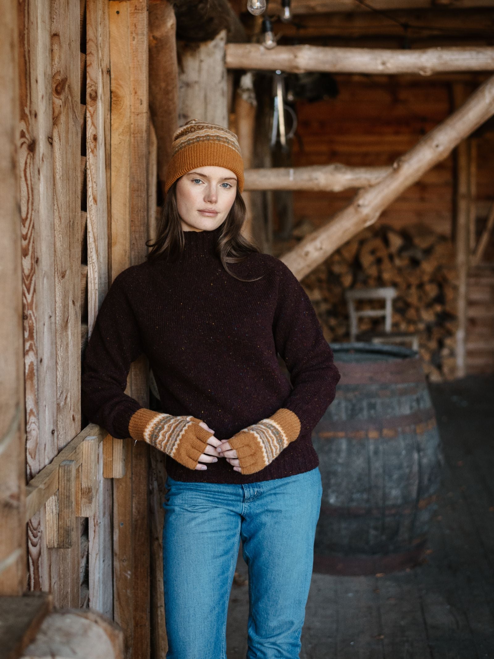 A woman wears the Campbell's of Beauly Lambswool Fairisle Hat, leaning against a wooden wall in a rustic cabin with stacked firewood and a barrel—showcasing cozy winter style.