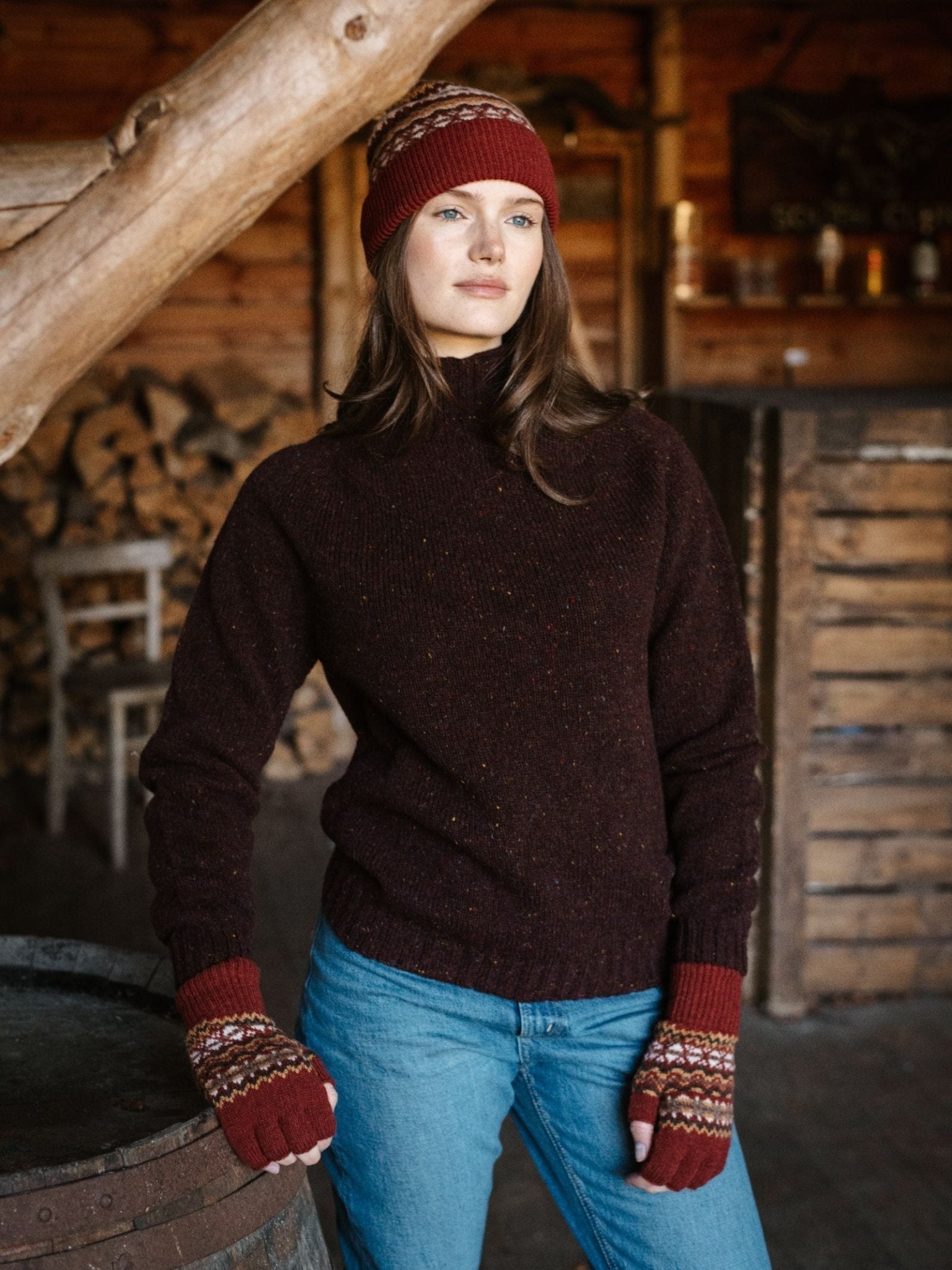 A woman in a maroon knit sweater, blue jeans, and Campbell's of Beauly Lambswool Fairisle Hat stands indoors by stacked firewood and rustic wooden walls, looking to the side—a true winter essential look.