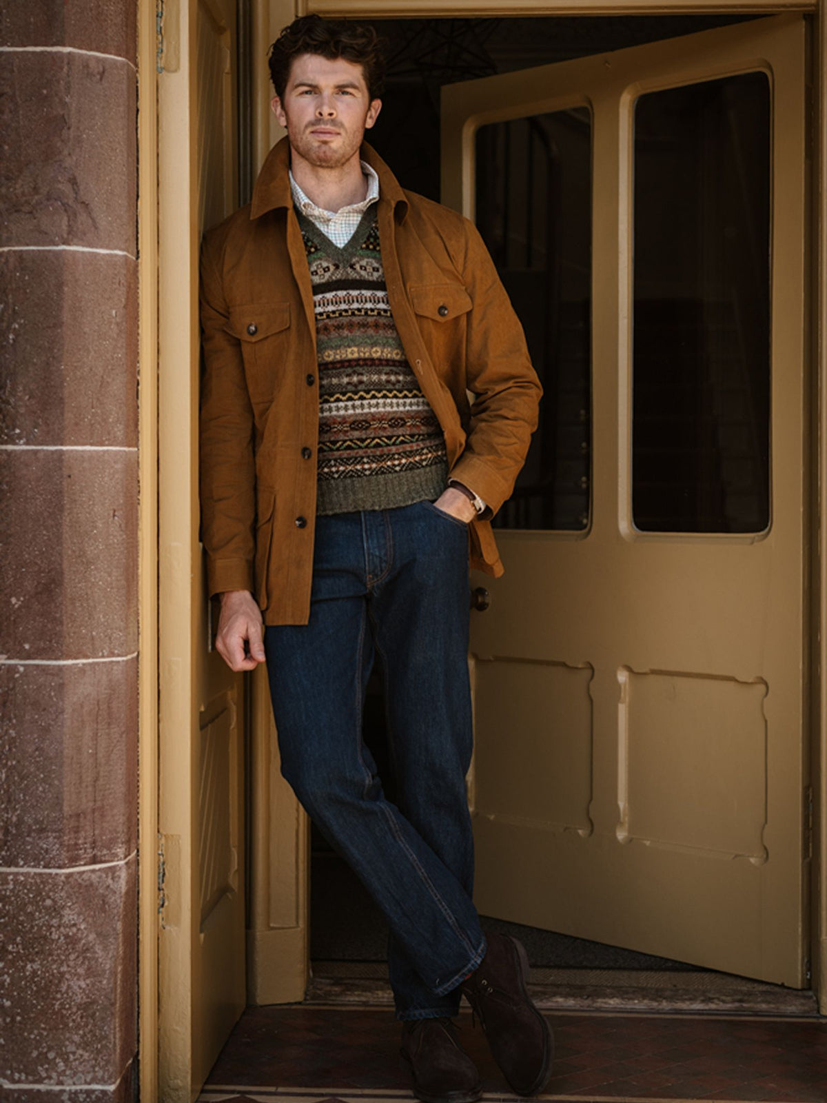 A man wearing a brown jacket, blue jeans, and dark shoes stands in a doorway, leaning against the doorframe with arms relaxed and one leg crossed over the other. Layered over his patterned sweater is the Campbell’s of Beauly Fairisle Slipover, a sleeveless Shetland wool piece that adds a touch of classic style to his look.