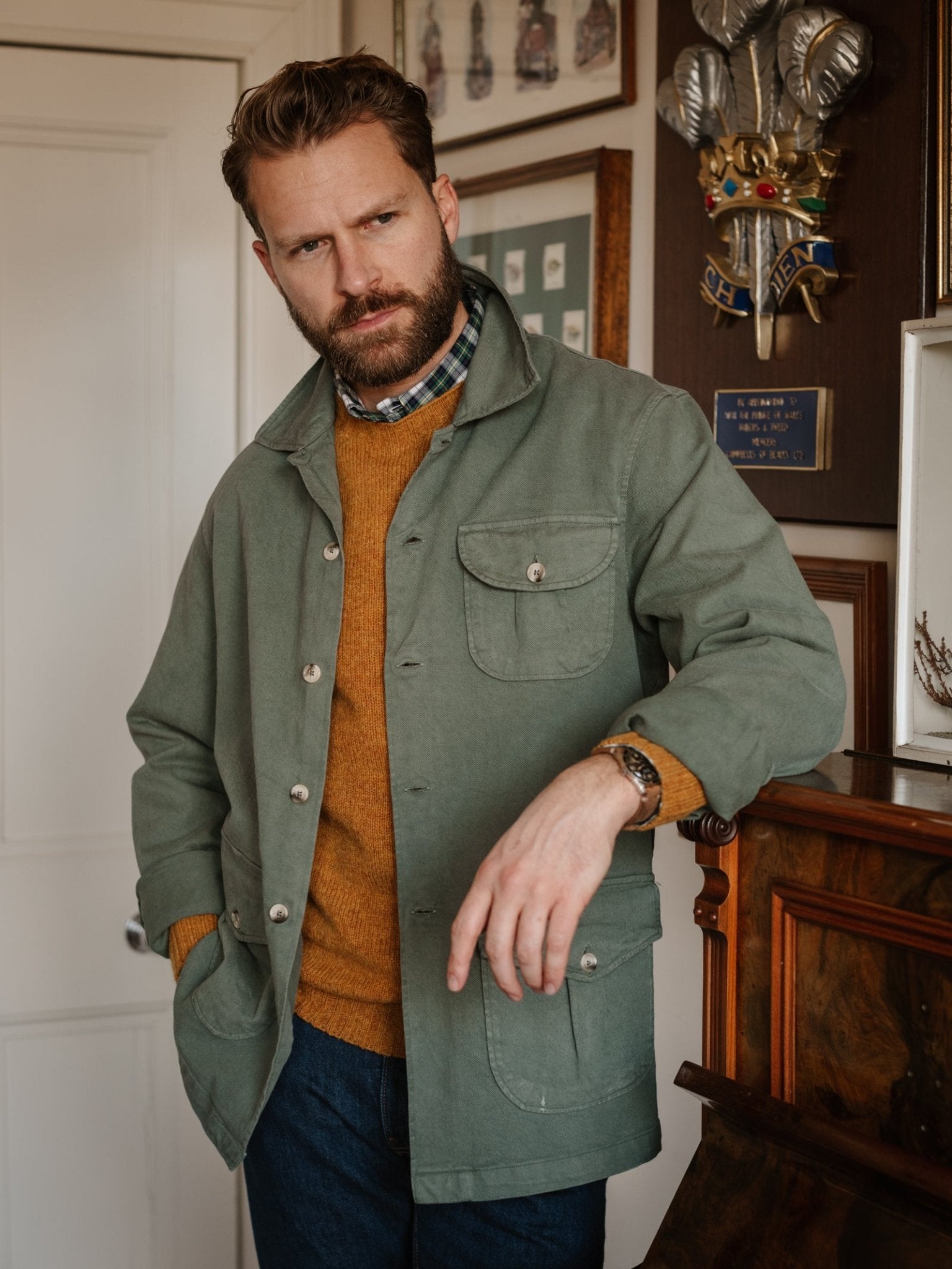 A bearded man wears a green Campbell’s of Beauly Field Chore Jacket, made in Europe from 100% cotton, over a mustard sweater and plaid shirt. He stands indoors by a wooden mantel, with framed pictures and a coat of arms behind him.