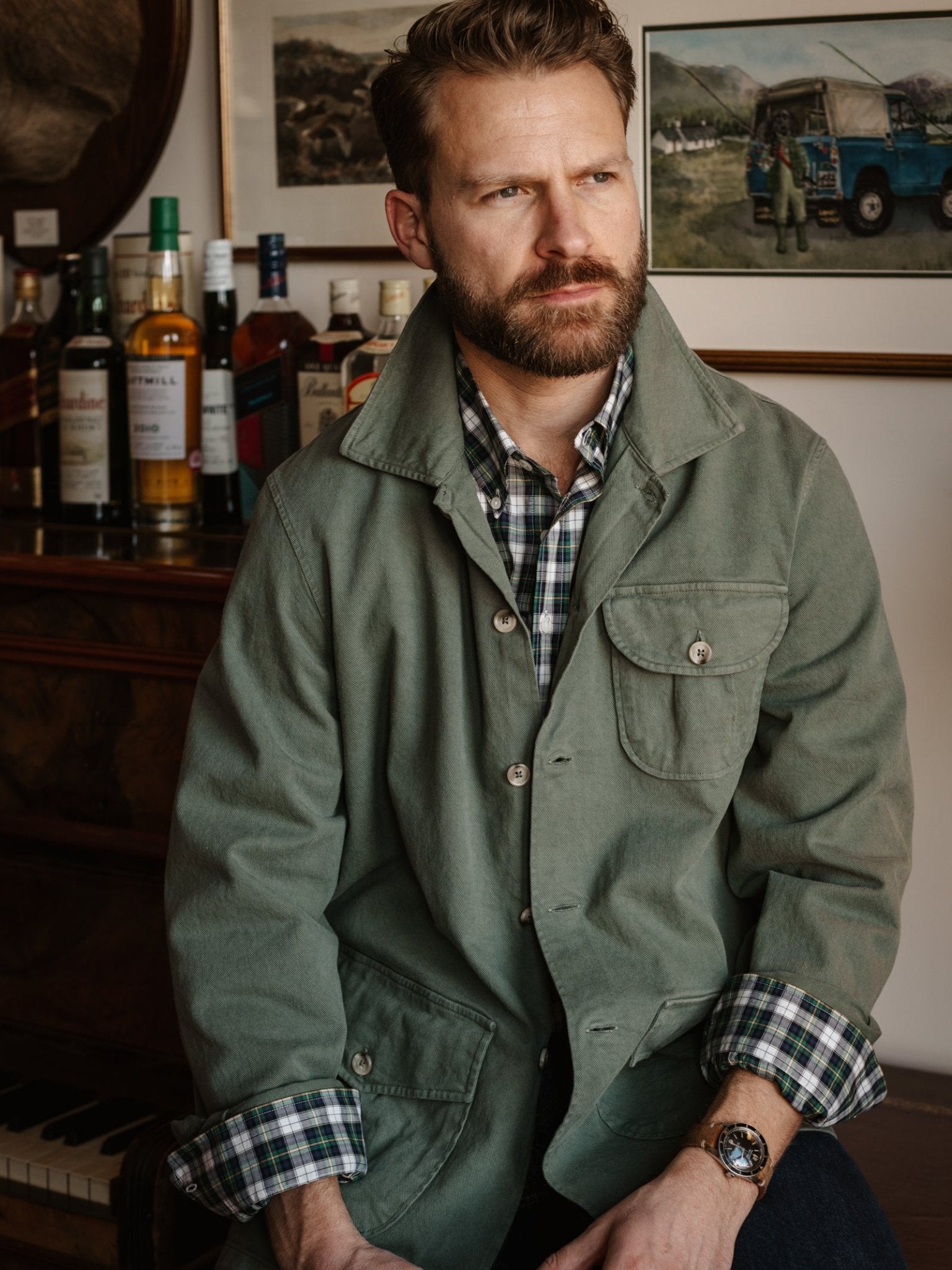 A bearded man wearing a Campbell's of Beauly Field Chore Jacket in Army, crafted from 100% cotton and made in Europe, sits indoors before a bar lined with bottles and framed art on the wall behind him.