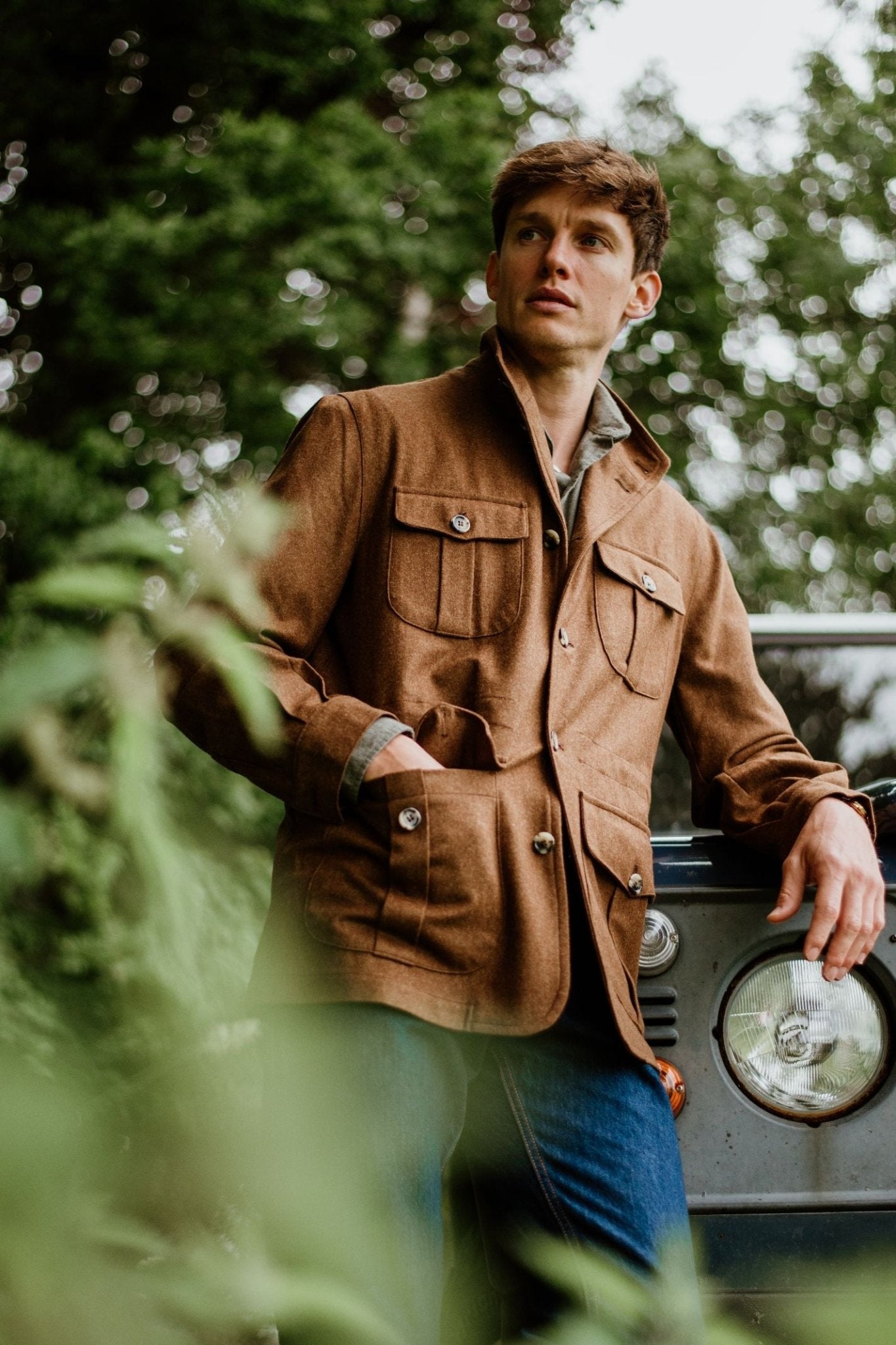 A man wearing the Campbells of Beauly Field Teba Jacket and blue jeans stands outdoors by a vintage vehicle, leaning on its front amid greenery and trees.