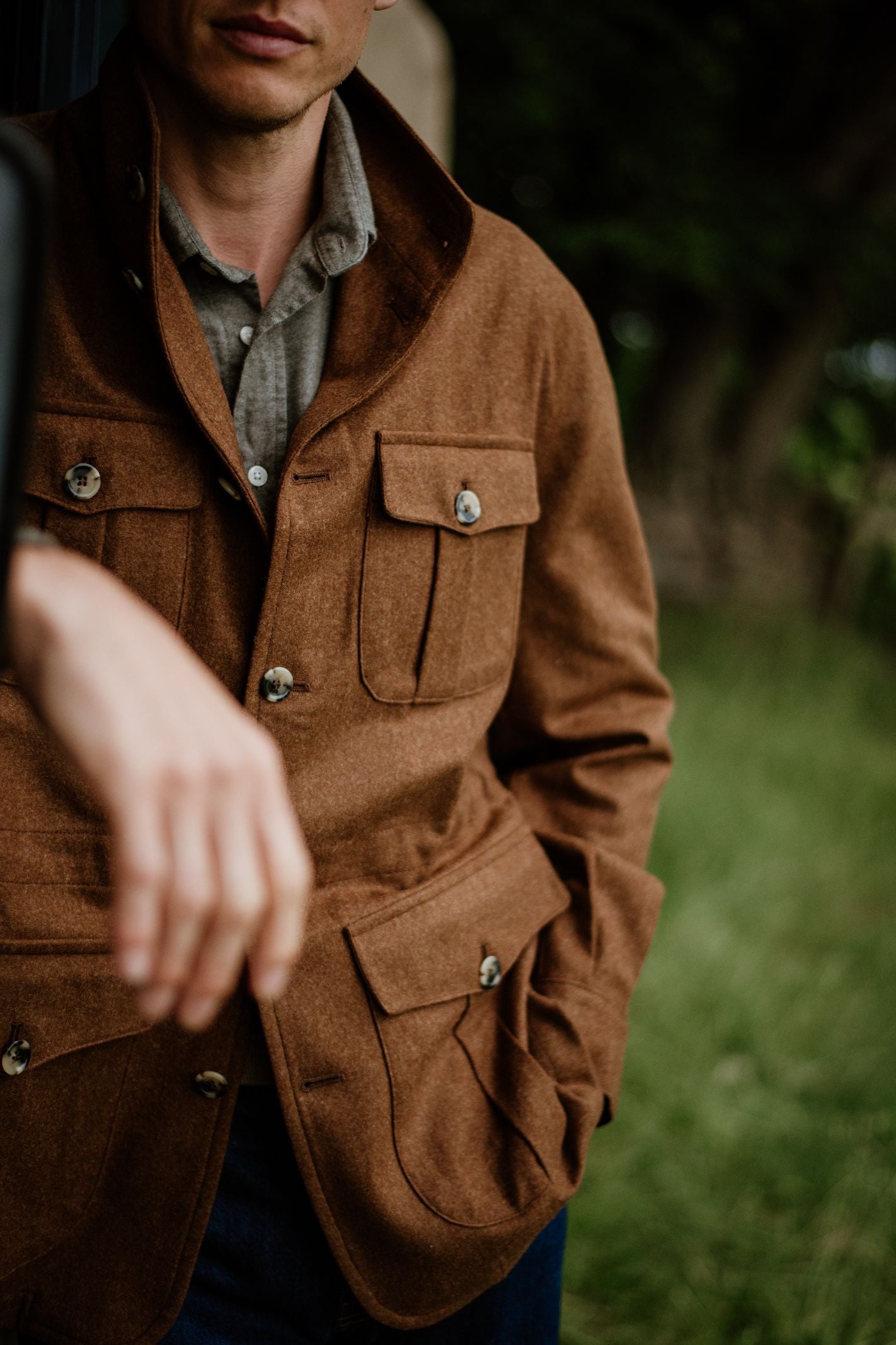 A man outdoors stands with one hand in his pocket, wearing a brown Campbell’s of Beauly Field Teba Jacket with large front pockets over a grey shirt, surrounded by green grass and trees.