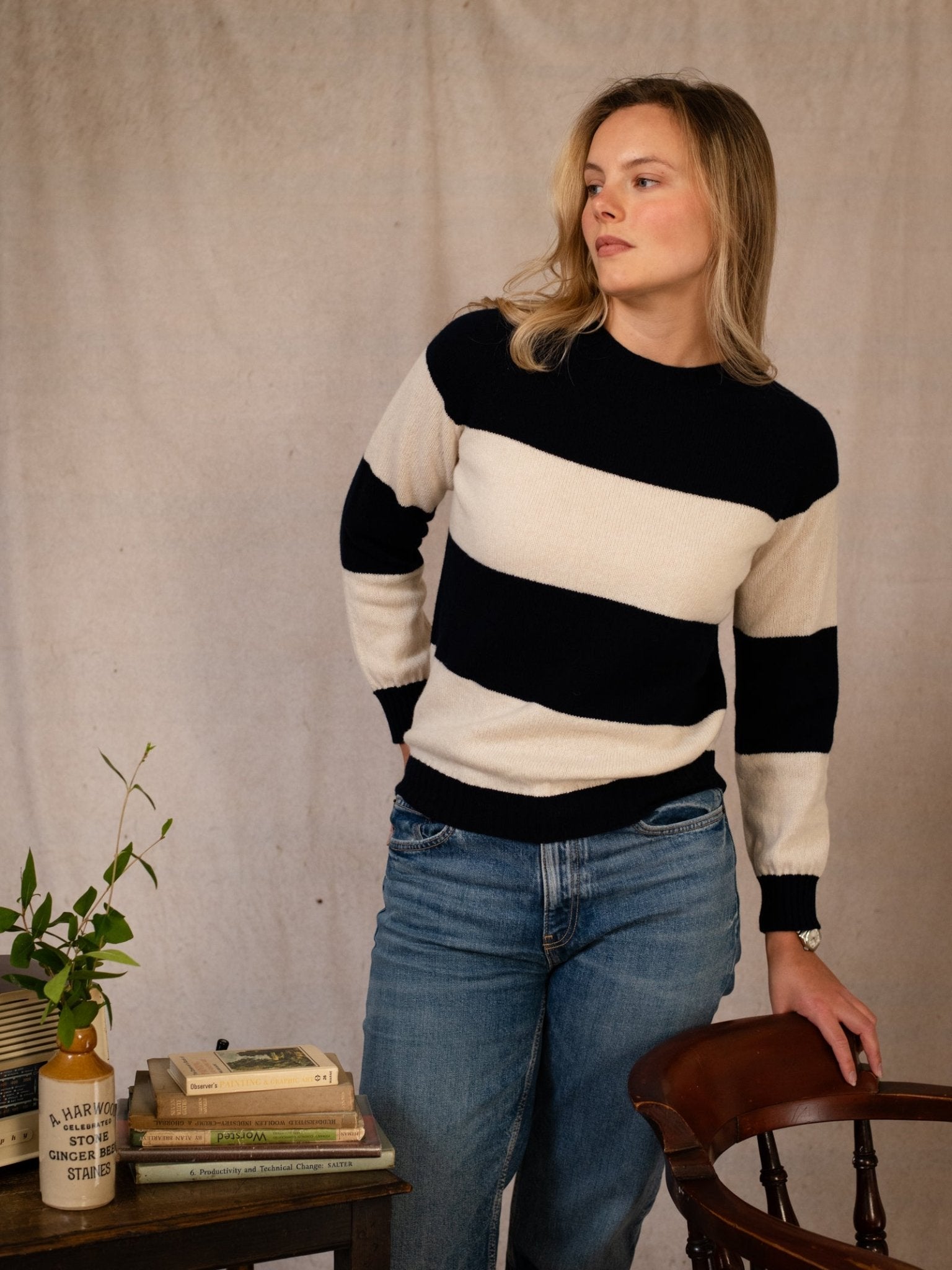 A woman wearing the Geelong Stripe Crew by Campbell's of Beauly—a relaxed fit, black and white striped crew neck jumper—stands with jeans beside a wooden chair and table, posing against a beige fabric backdrop. Expertly knitted in Scotland.
