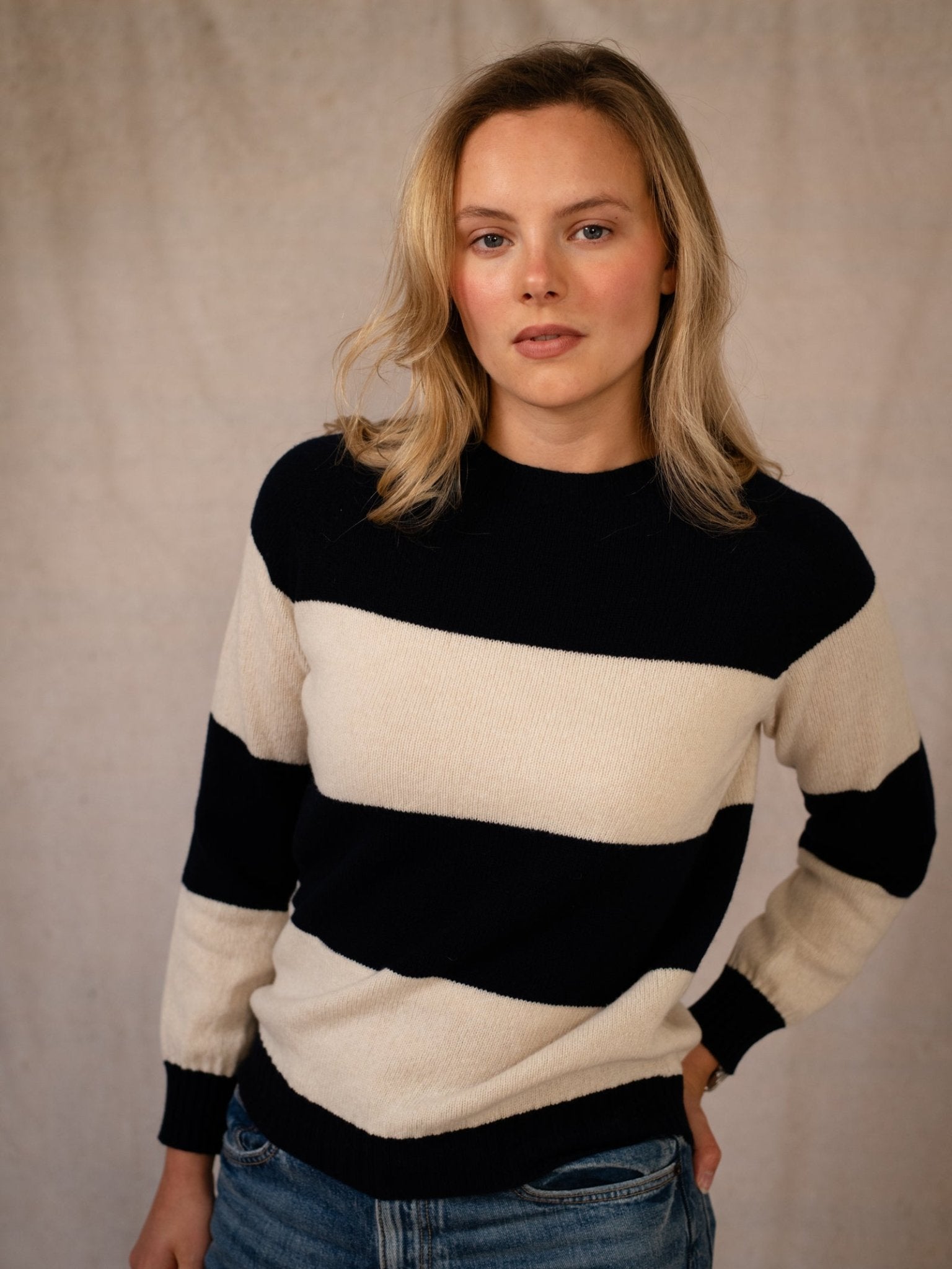 A woman with blonde hair wears the Campbell's of Beauly Geelong Stripe Crew in navy and blue jeans, standing against a neutral beige background and looking at the camera with a calm expression.
