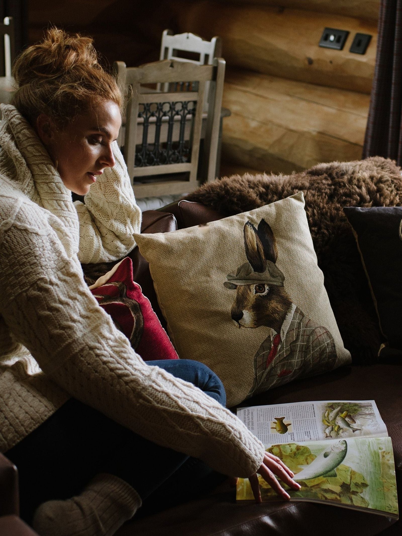 A woman in a cozy sweater sits on a couch, looking at an open book or magazine. Beside her is the Dapper Hare Cushion Cover by Campbell’s of Beauly, featuring a charming rabbit illustration dressed in clothes. The setting appears warm and rustic.