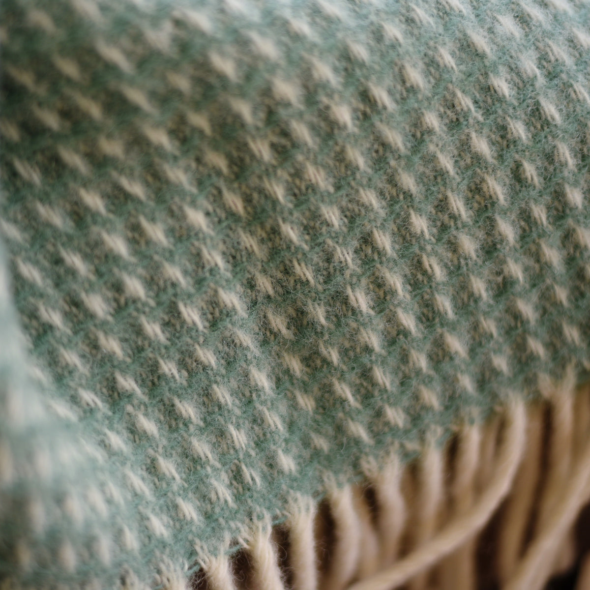 Close-up of a soft woven fabric featuring a light green and cream houndstooth pattern with beige fringe at the edge, from the Honeycomb Lambswool Throw by Campbells of Beauly.