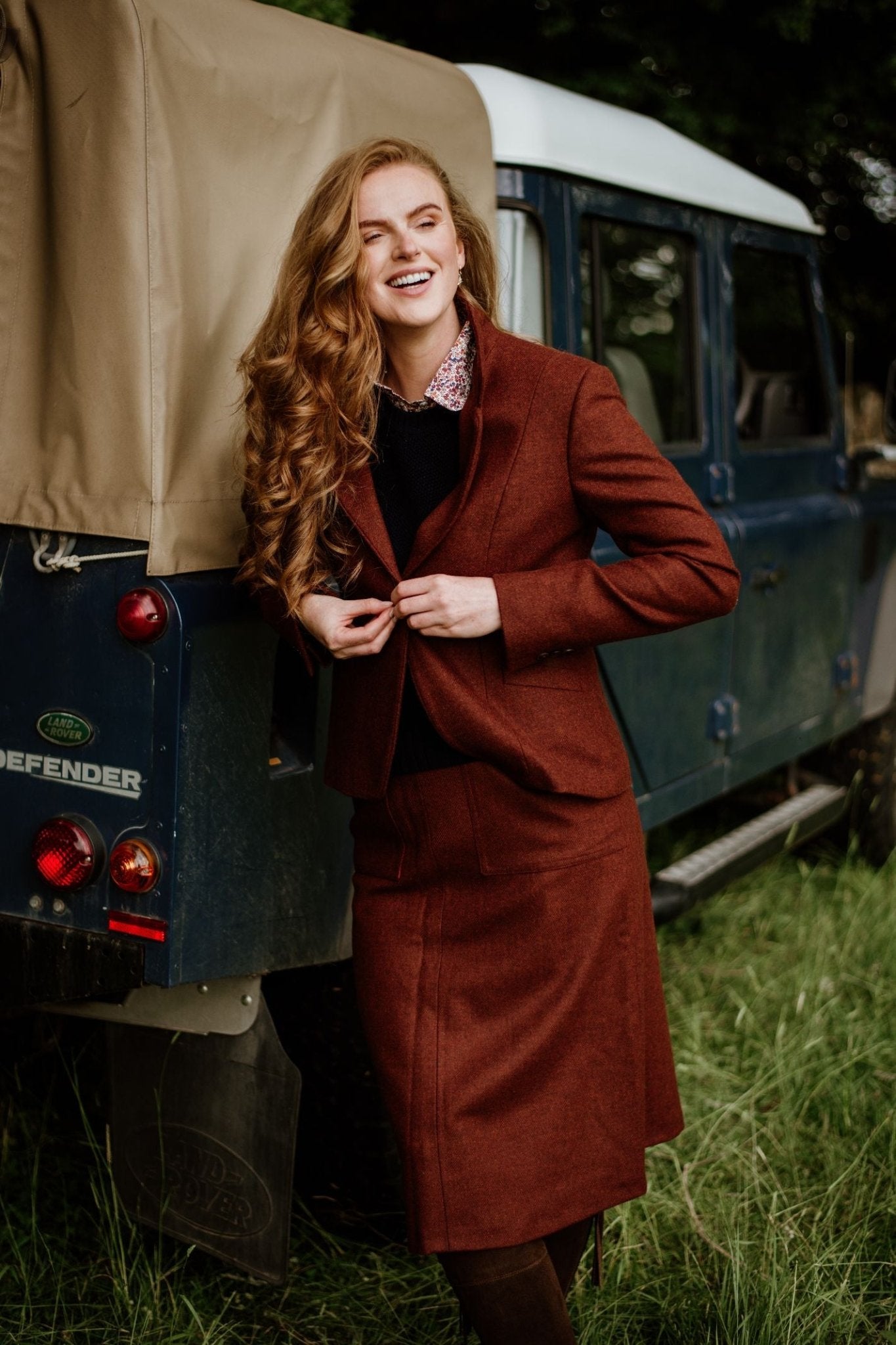 A woman with long red hair, smiling in a House Jacket by Campbells of Beauly and a reddish-brown skirt suit, stands beside a blue Land Rover Defender on grass with trees in the background.