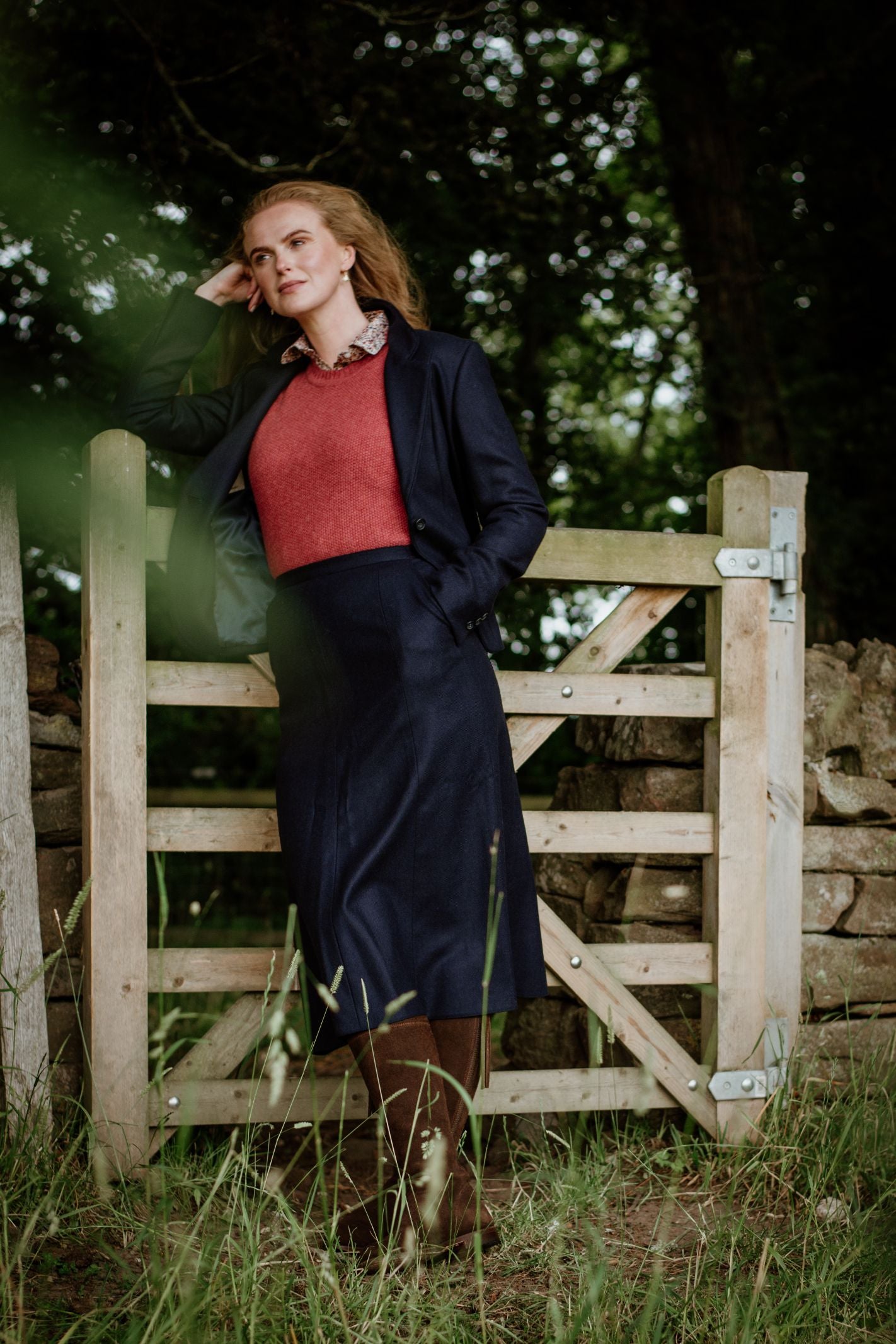 Wearing the Campbells of Beauly House Jacket, a woman with long hair stands by a wooden gate in nature, her head resting on her hand as she gazes thoughtfully into the distance amid tall grass and trees.