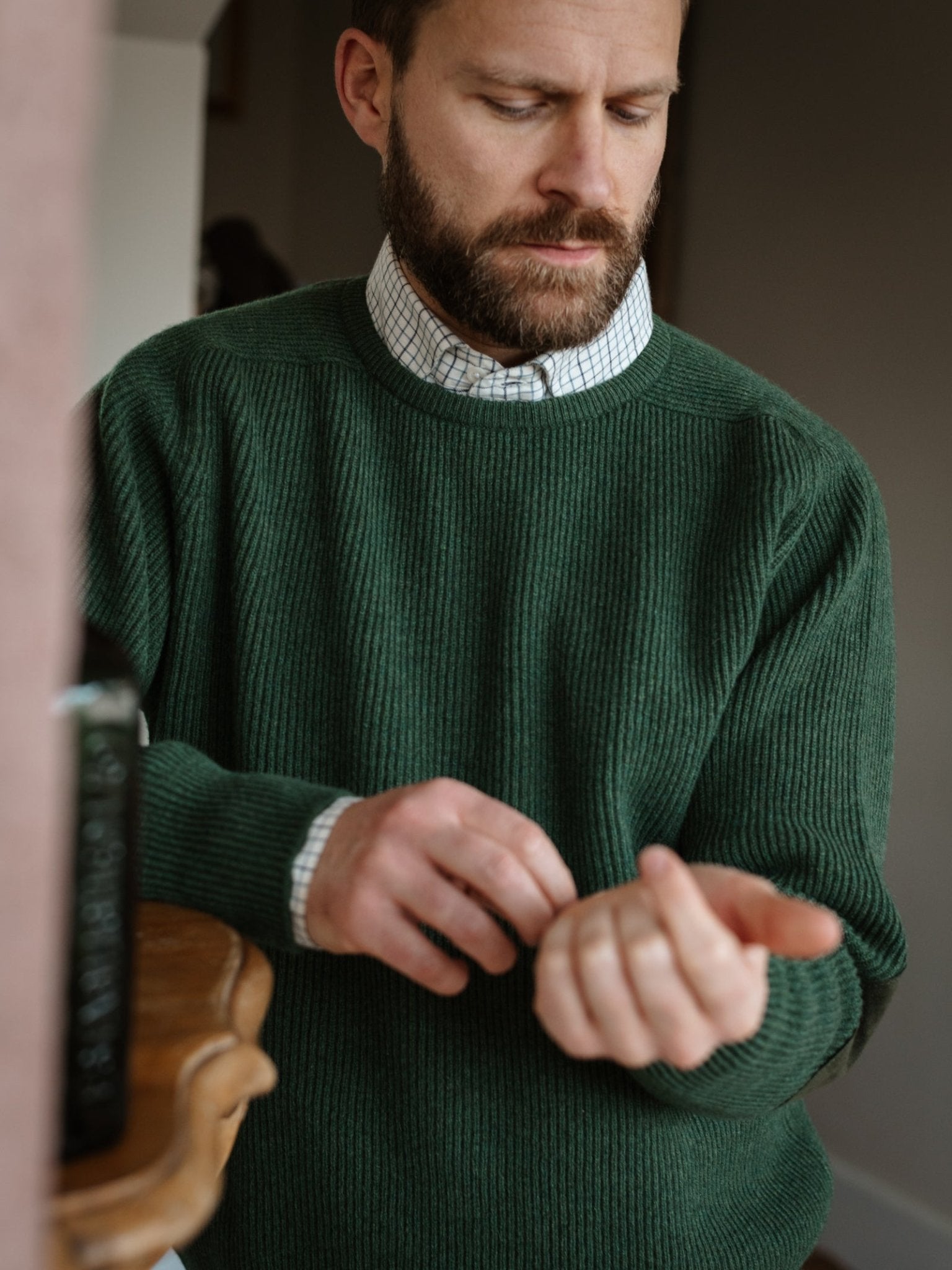 A bearded man wears the Campbell's of Beauly Fisherman's Rib Crew Jumper in green over a checked shirt, adjusting his cuff indoors by a wooden surface, highlighting classic Scottish knitwear style.