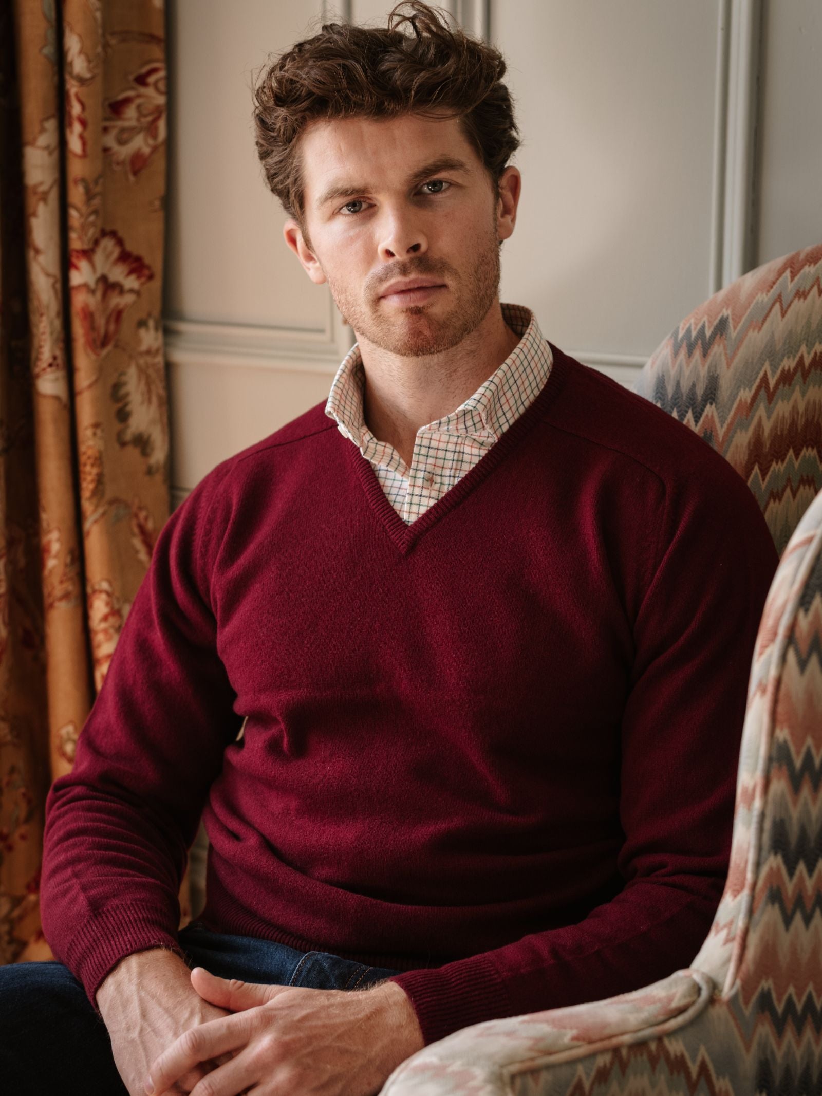 A man with short, wavy brown hair and light stubble sits in an upholstered armchair, wearing a Campbell's of Beauly Lambswool V-Neck Jumper over a checked shirt. Floral curtains and paneled walls complete the cozy scene.