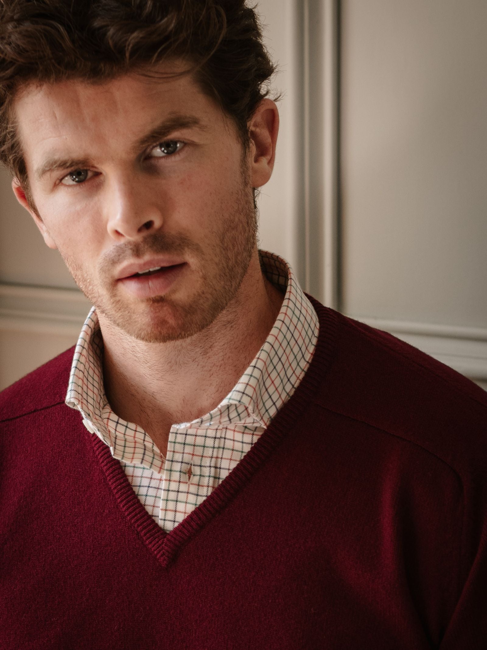 A man with short brown hair and light stubble wears a Campbell's of Beauly Lambswool V-Neck Jumper over a checked shirt, standing indoors against a neutral wall and looking directly at the camera with a neutral expression.