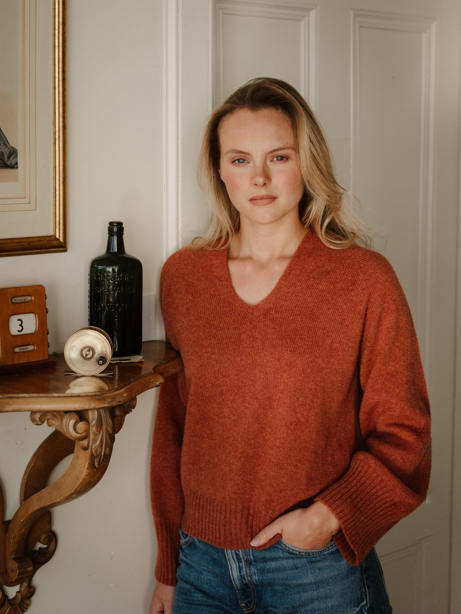 A blonde woman wears the Campbell's of Beauly Geelong Lambswool V Jumper in copper with blue jeans, standing indoors by a wooden shelf and white paneled wall, calmly looking at the camera with one hand in her pocket.