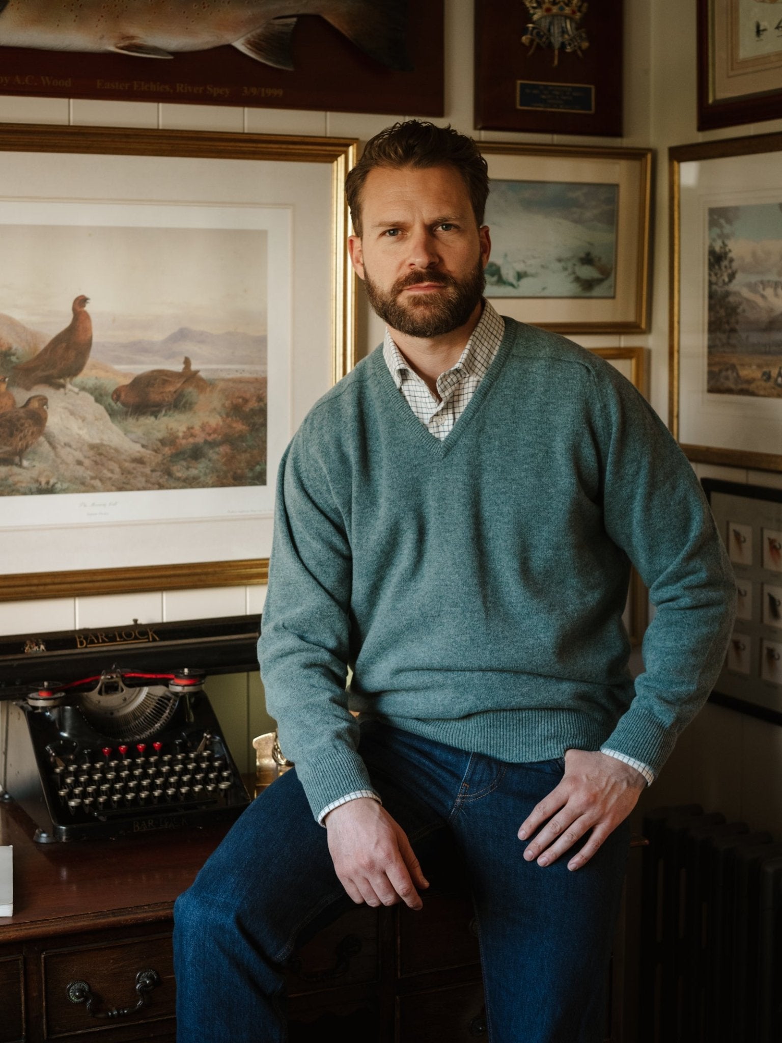A bearded man sits at a desk wearing the Campbell's of Beauly Lambswool V-Neck Jumper over a checkered shirt and blue jeans, surrounded by framed artwork and a vintage typewriter.