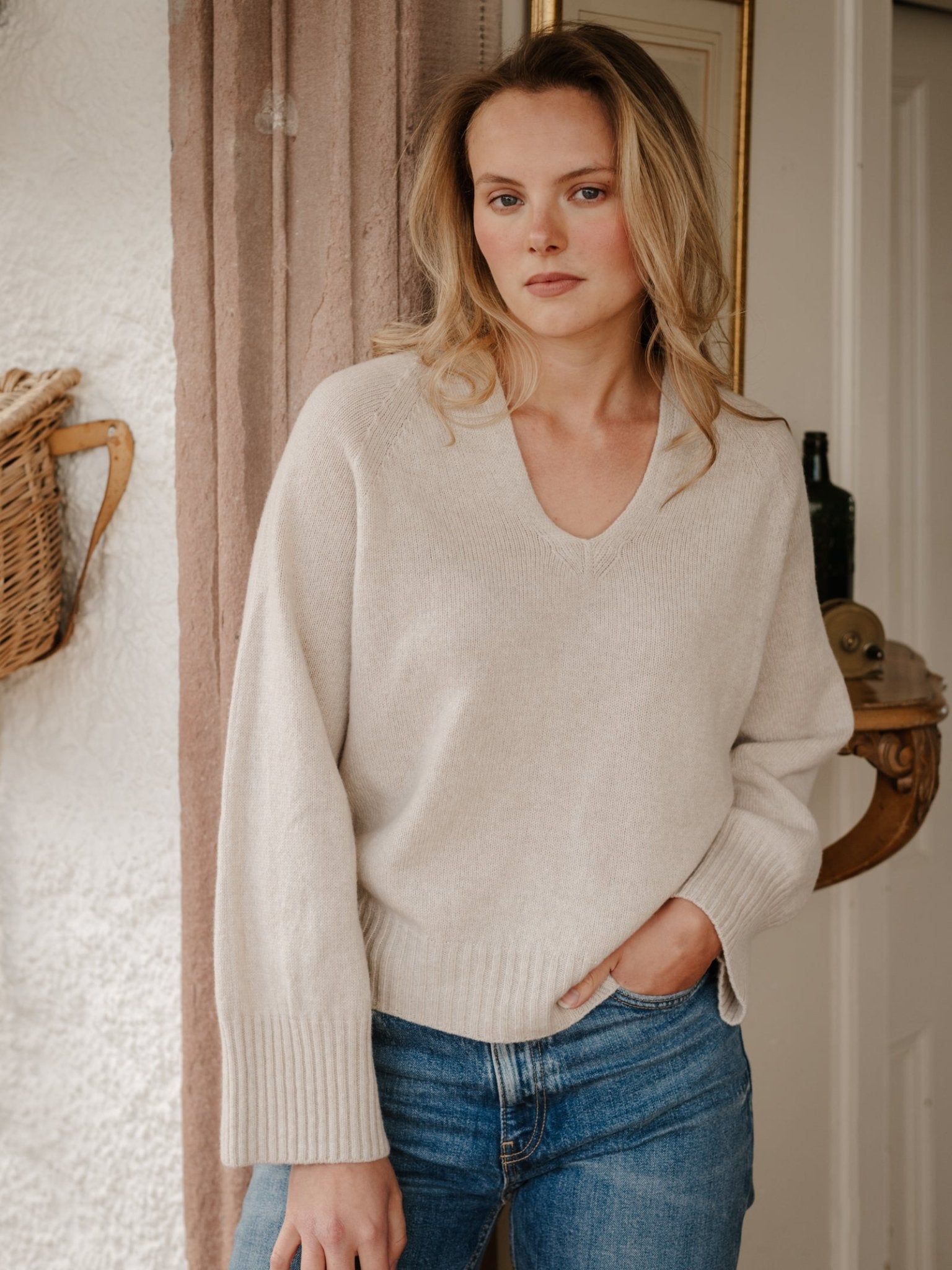 A woman with blonde hair stands indoors, one hand in her pocket, wearing the Campbell's of Beauly Geelong Lambswool V Jumper in Natural and blue jeans. She has a neutral expression and poses before a curtain and textured wall with a hanging basket.