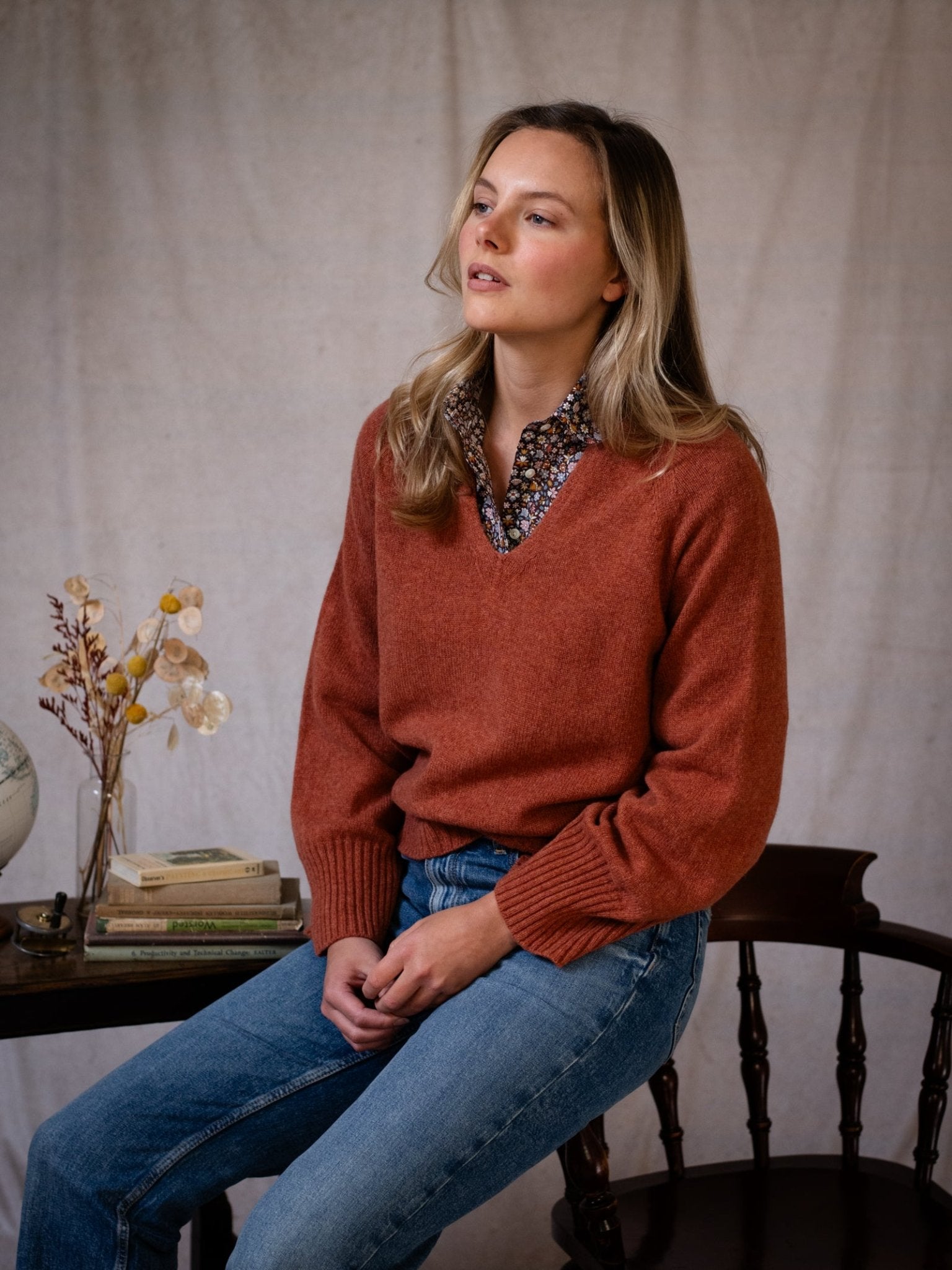 A woman with long blonde hair wears the Campbell's of Beauly Geelong Lambswool V Jumper in rust over a floral blouse and blue jeans, seated beside a table with books, dried flowers, and a globe against a neutral fabric backdrop.