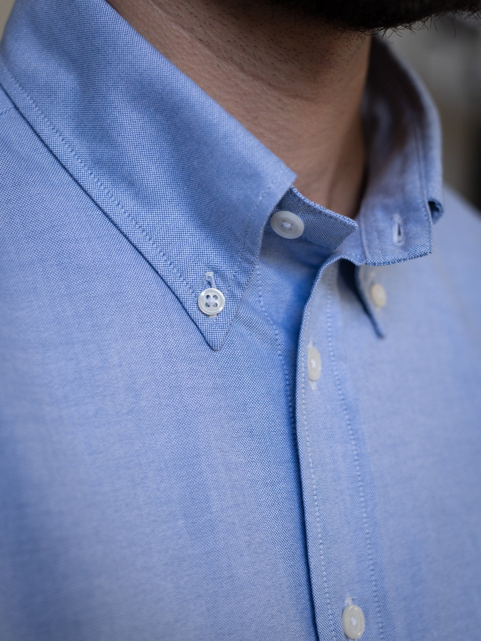 Close-up of a person wearing the Campbell's of Beauly Oxford Button Down Shirt in light blue cotton, featuring a button-down collar with white buttons and part of the neck and jawline with a light beard visible.