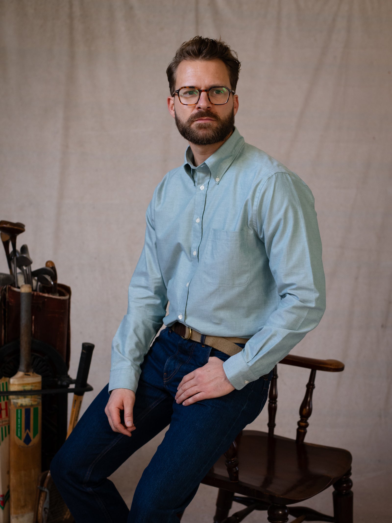 A bearded man in glasses wears a light green Oxford Button Down Shirt by Campbell’s of Beauly and dark jeans as he sits on a wooden chair, with vintage sports equipment like cricket bats displayed beside him.