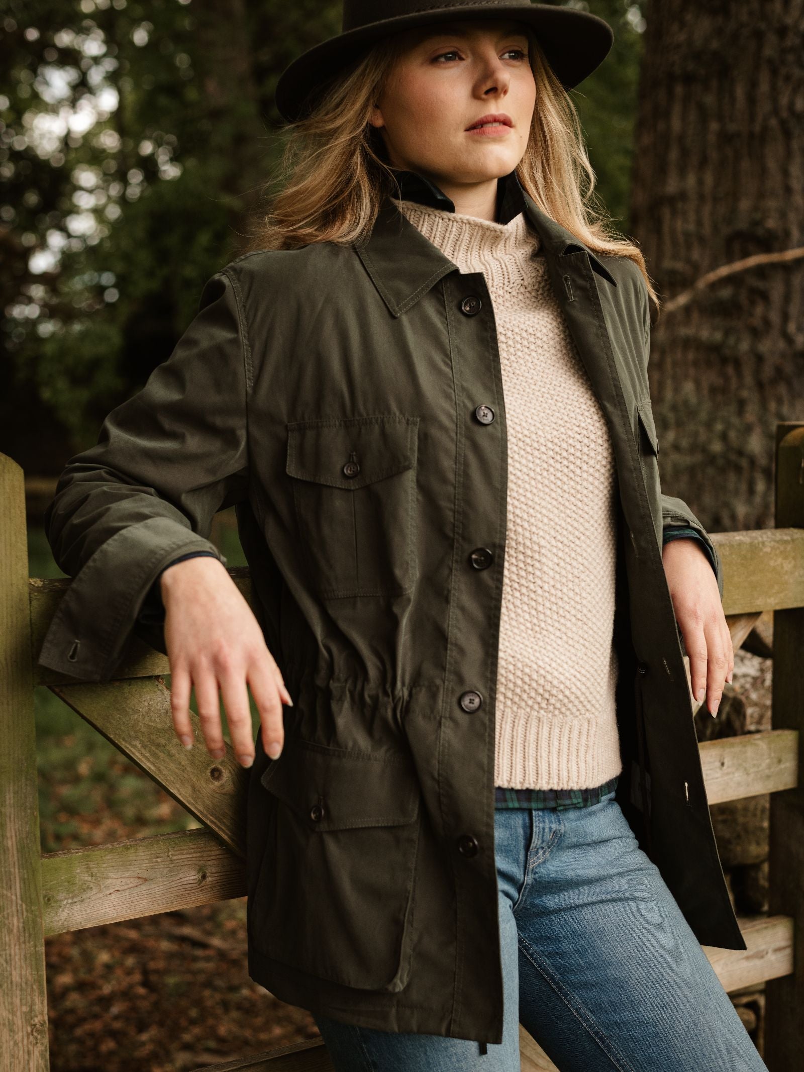 A woman in a Campbell’s of Beauly Lightweight Aigas Field Jacket, cream knit sweater, blue jeans, and wide-brimmed hat leans against a wooden fence outdoors, trees visible in the background.