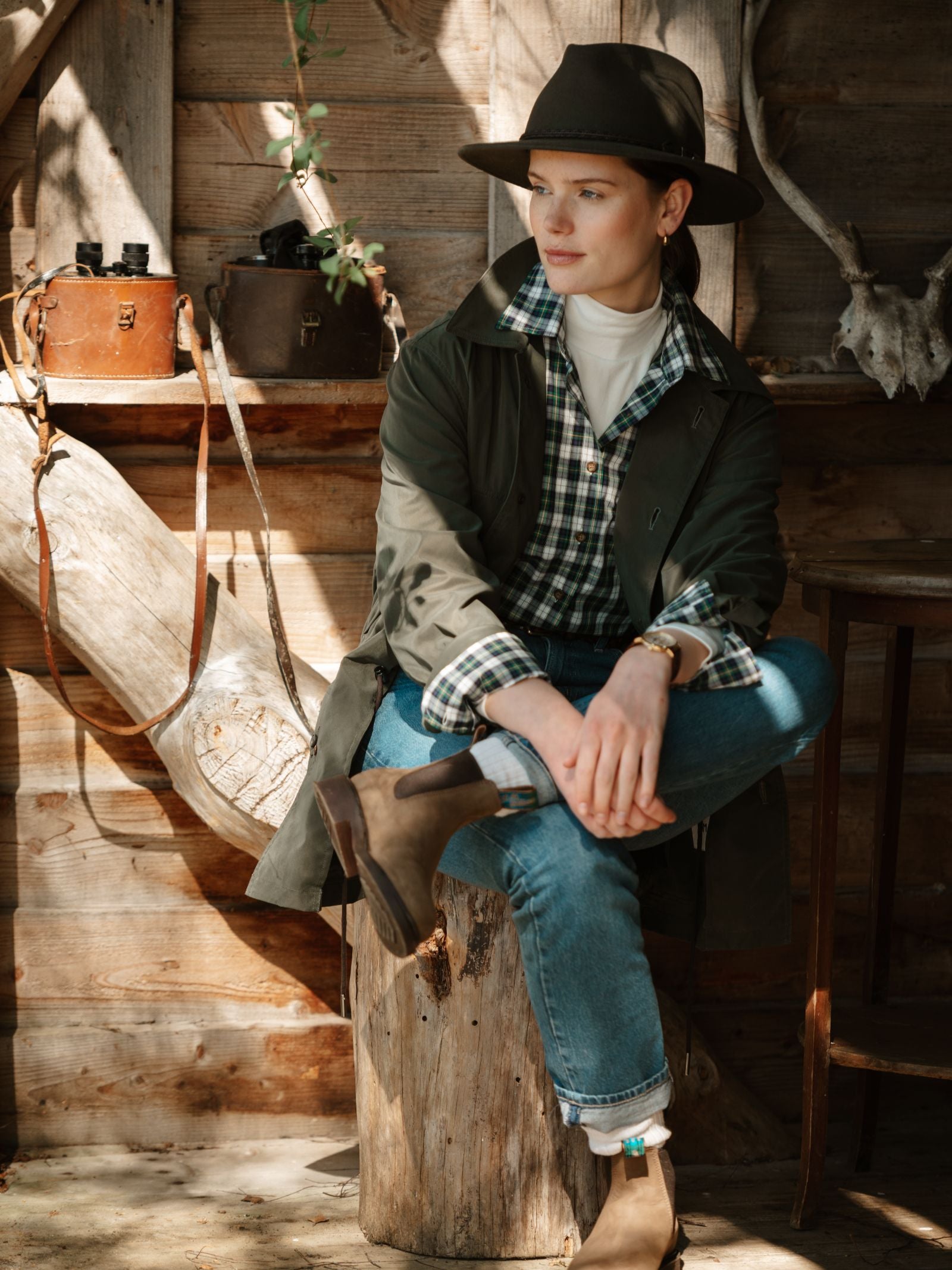 A woman in a hat, plaid shirt, Campbell's of Beauly Lightweight Aigas Field Jacket, jeans, and boots sits on a tree stump indoors with sunlight streaming in amid vintage bags and antlers against a rustic wooden backdrop.