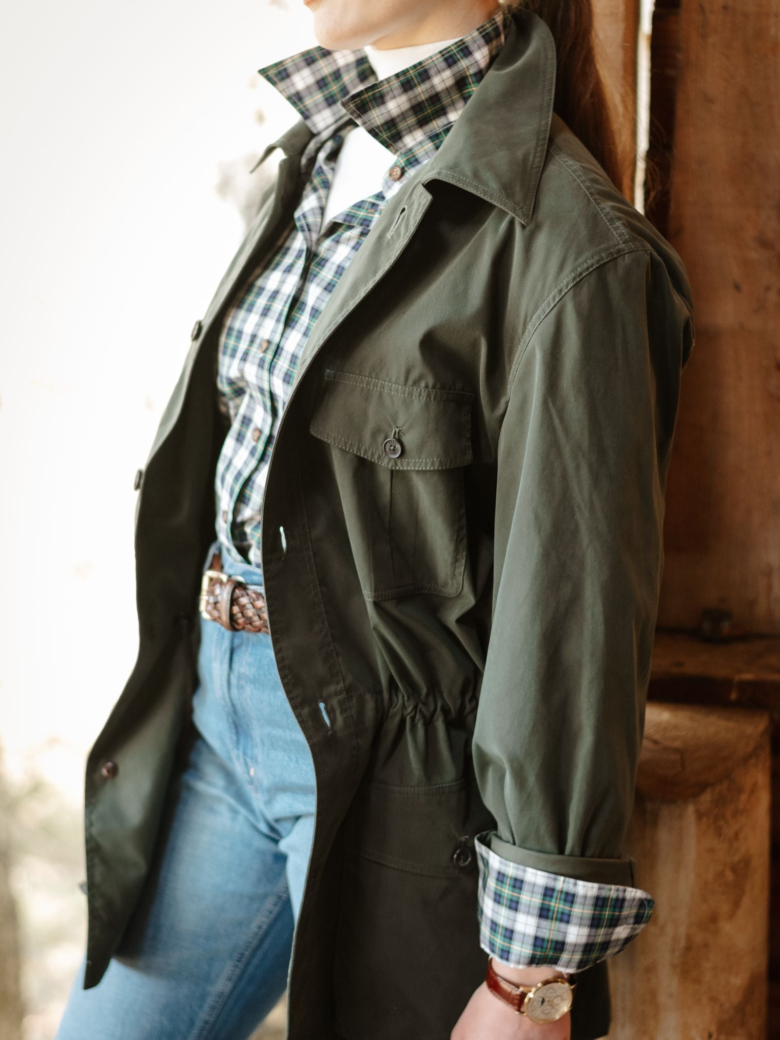 A person stands by a wooden post, wearing Campbell's of Beauly Lightweight Aigas Field Jacket over a plaid shirt and white tee, paired with blue jeans and a brown belt. Their face is partially out of frame.