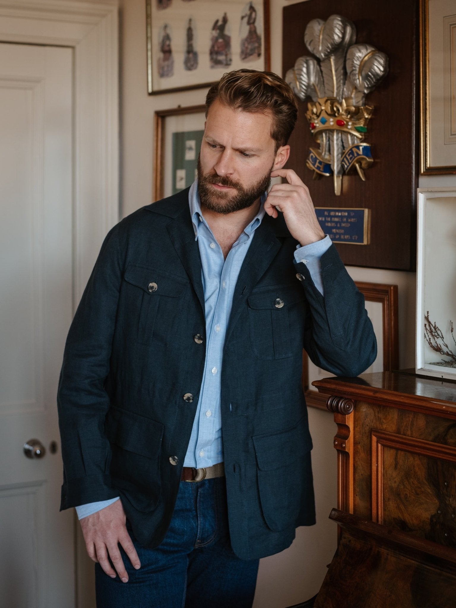 A bearded man wears a Campbell's of Beauly Navy Linen Safari Jacket over a light blue shirt and jeans, standing indoors amid framed art and wooden decor, touching his ear with a thoughtful look.