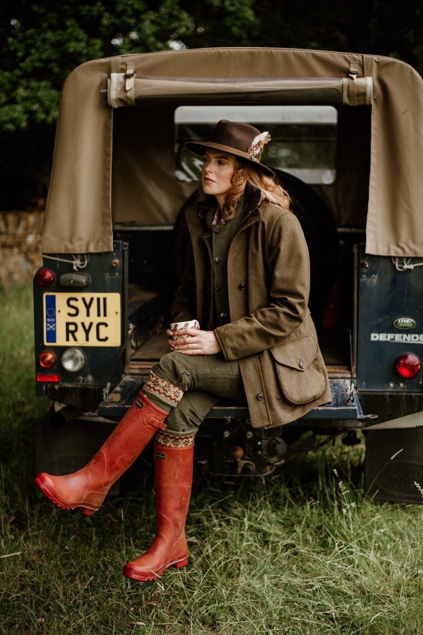 A woman in the Campbells of Beauly Loden Green Field Coat and red boots sits on a vintage Land Rover Defender, holding a mug. Amid grass and trees, she highlights this classic outerwear crafted from Austrian loden fabric.