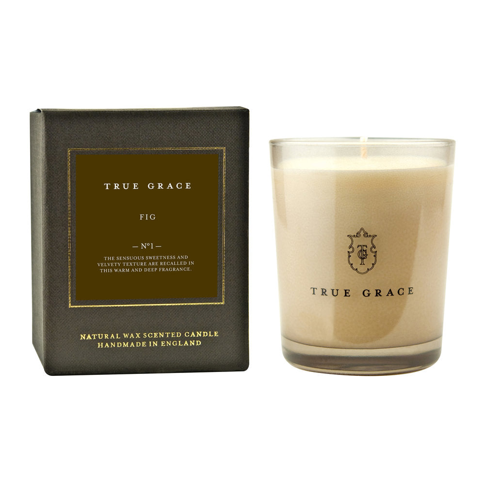 A True Grace Fig Candle with cream wax in a clear glass holder sits beside its black and gold box, labeled True Grace Fig and noting its handmade in England from natural wax.