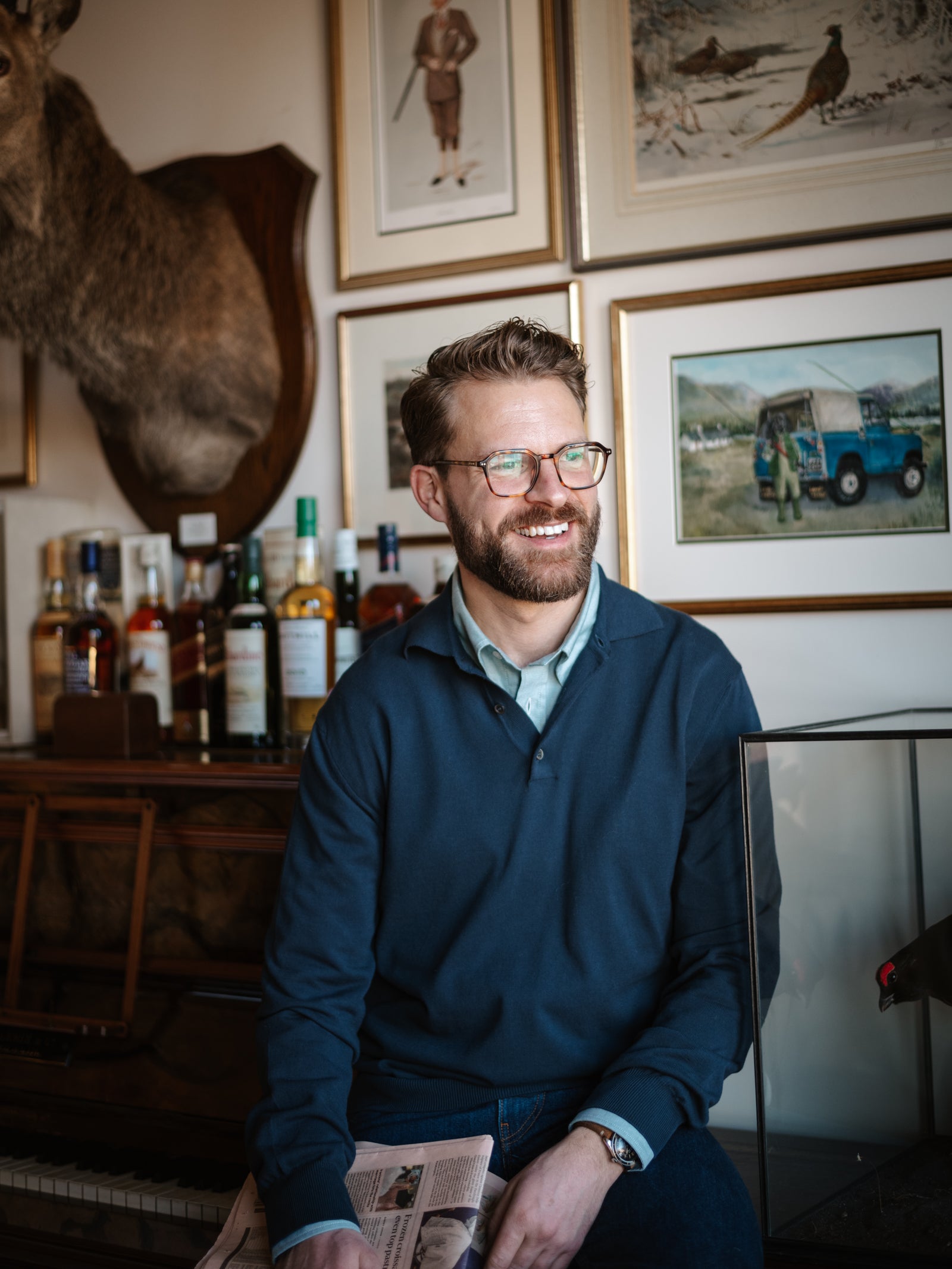 A smiling bearded man with glasses wears a Campbell's of Beauly Cotton Rugby Shirt as he sits in a wood-panelled room decorated with hunting prints, a deer mount, and whisky bottles—a perfect spot for summer layering.