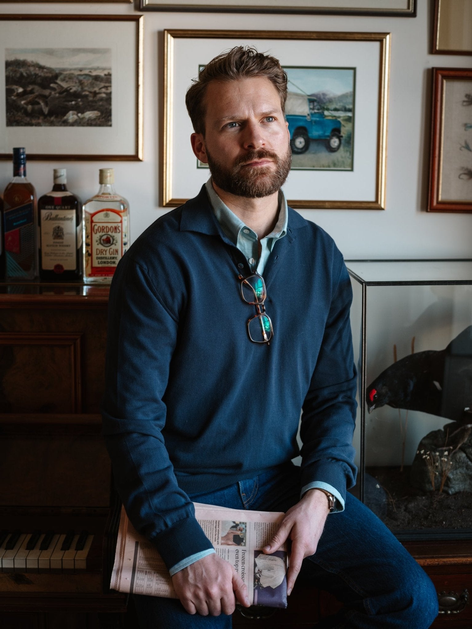 A man in a navy Campbell's of Beauly Cotton Rugby, ideal for summer layering, sits on a piano bench with a newspaper. Glasses hang from his shirt; behind him are bottles, framed pictures, and part of a taxidermy bird.