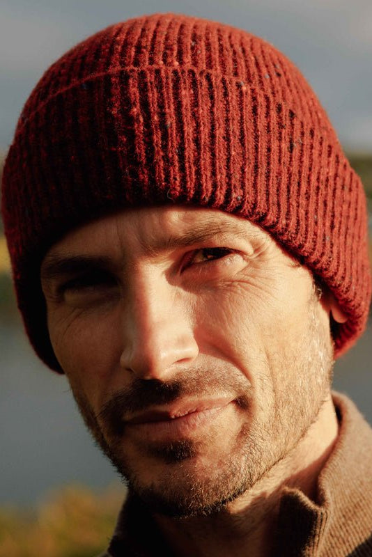 A man stands outdoors in soft sunlight, wearing the Campbells of Beauly Donegal Lambswool Hat—a red knit beanie crafted from Donegal yarns—with a brown jacket and a blurred backdrop of water and greenery.