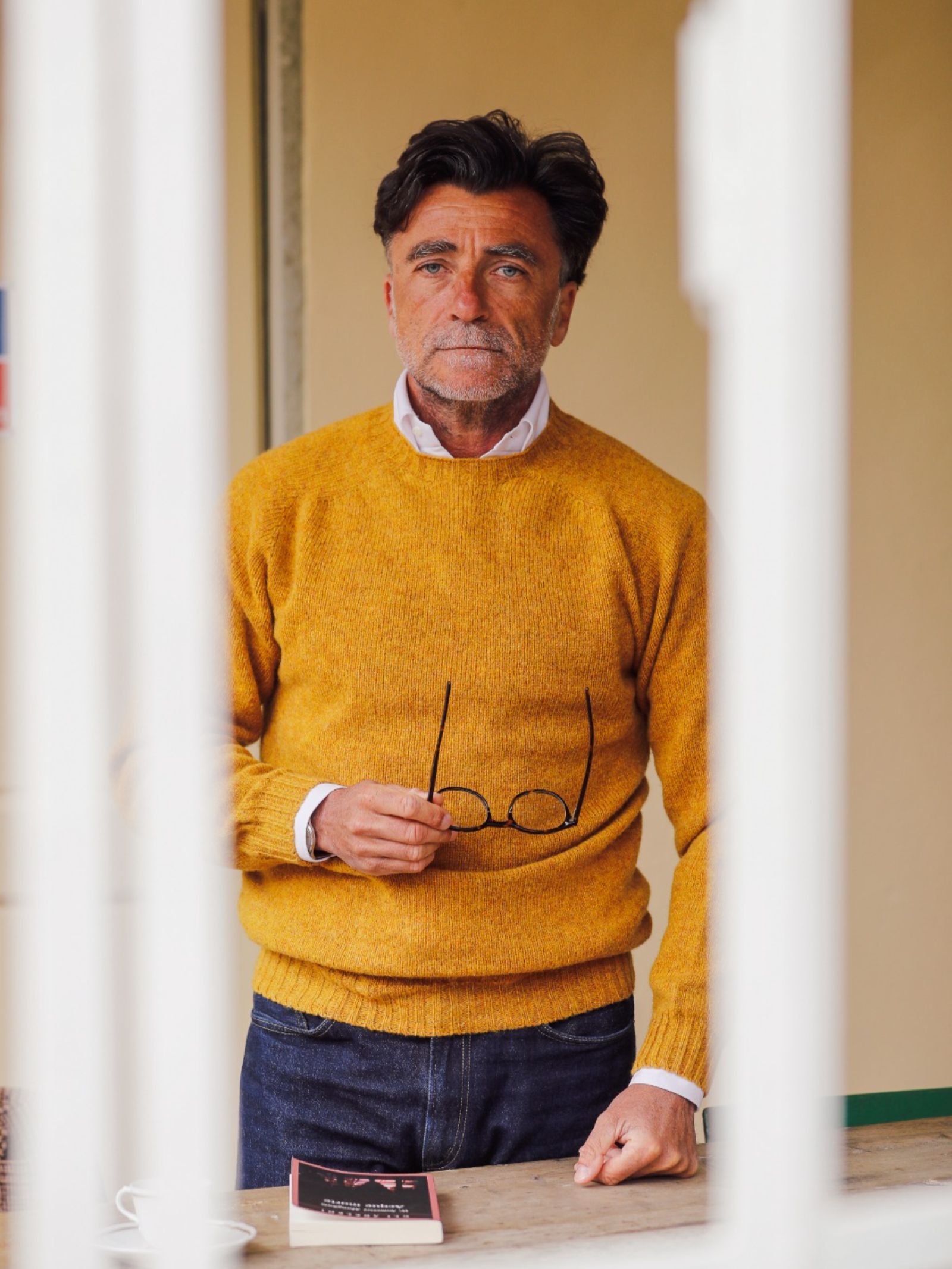 A middle-aged man with dark hair and a beard, dressed in a mustard yellow Campbell's of Beauly Shetland Jumper and jeans, stands indoors holding eyeglasses. A book and cup rest on the table before him.